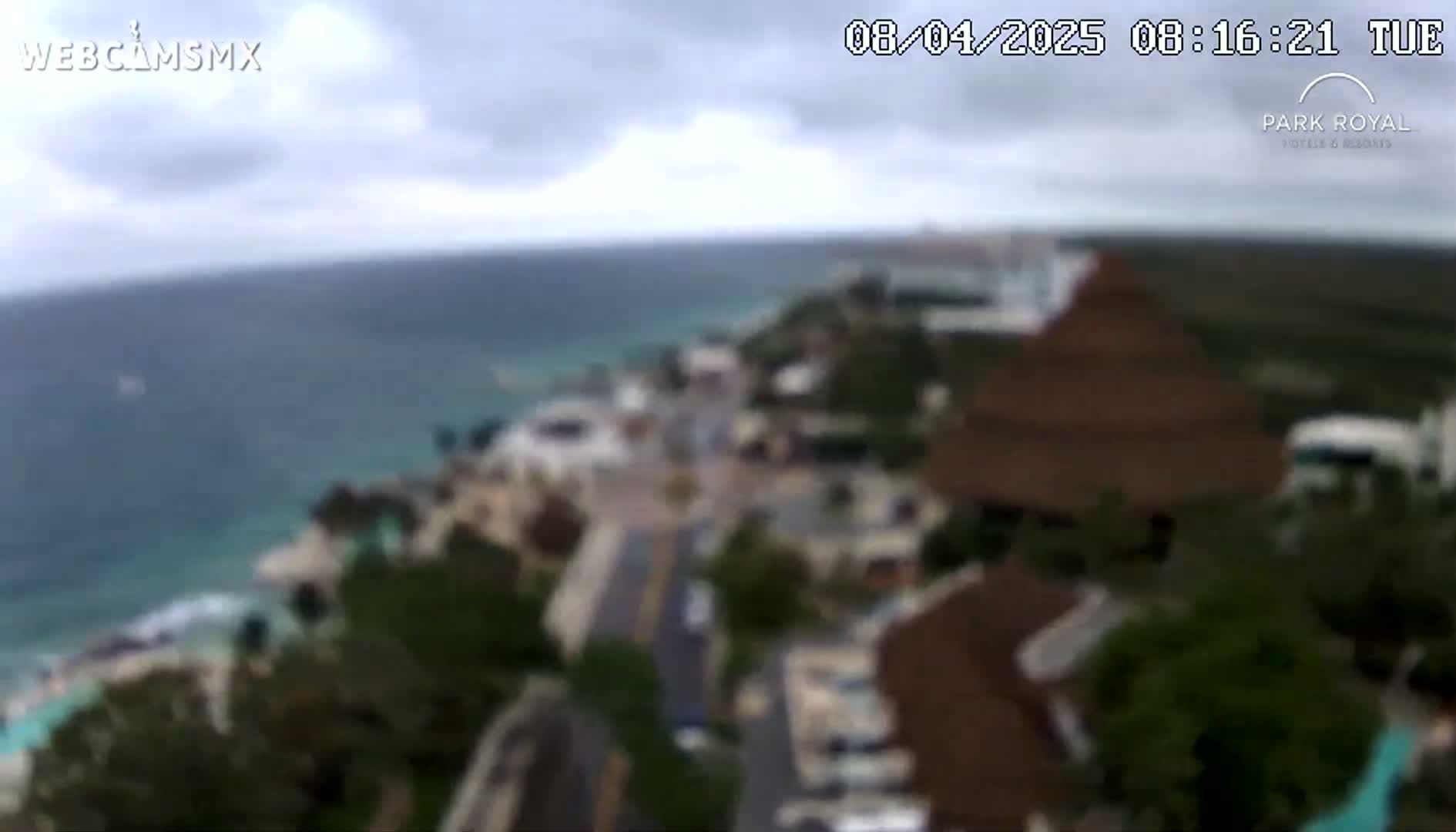 A blurry, high-angle view shows a coastal town under overcast skies, with a body of calm water in the background.