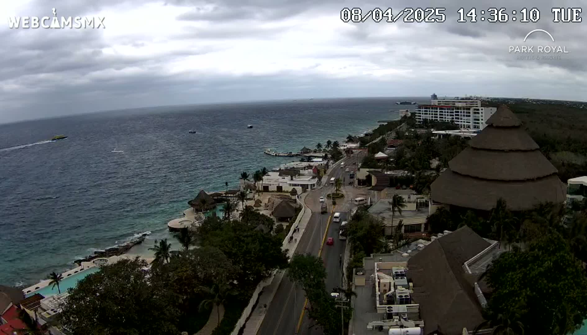 An aerial view of a coastal resort town on an overcast day shows a road leading to buildings and a beach with turquoise water, and boats in the distance.