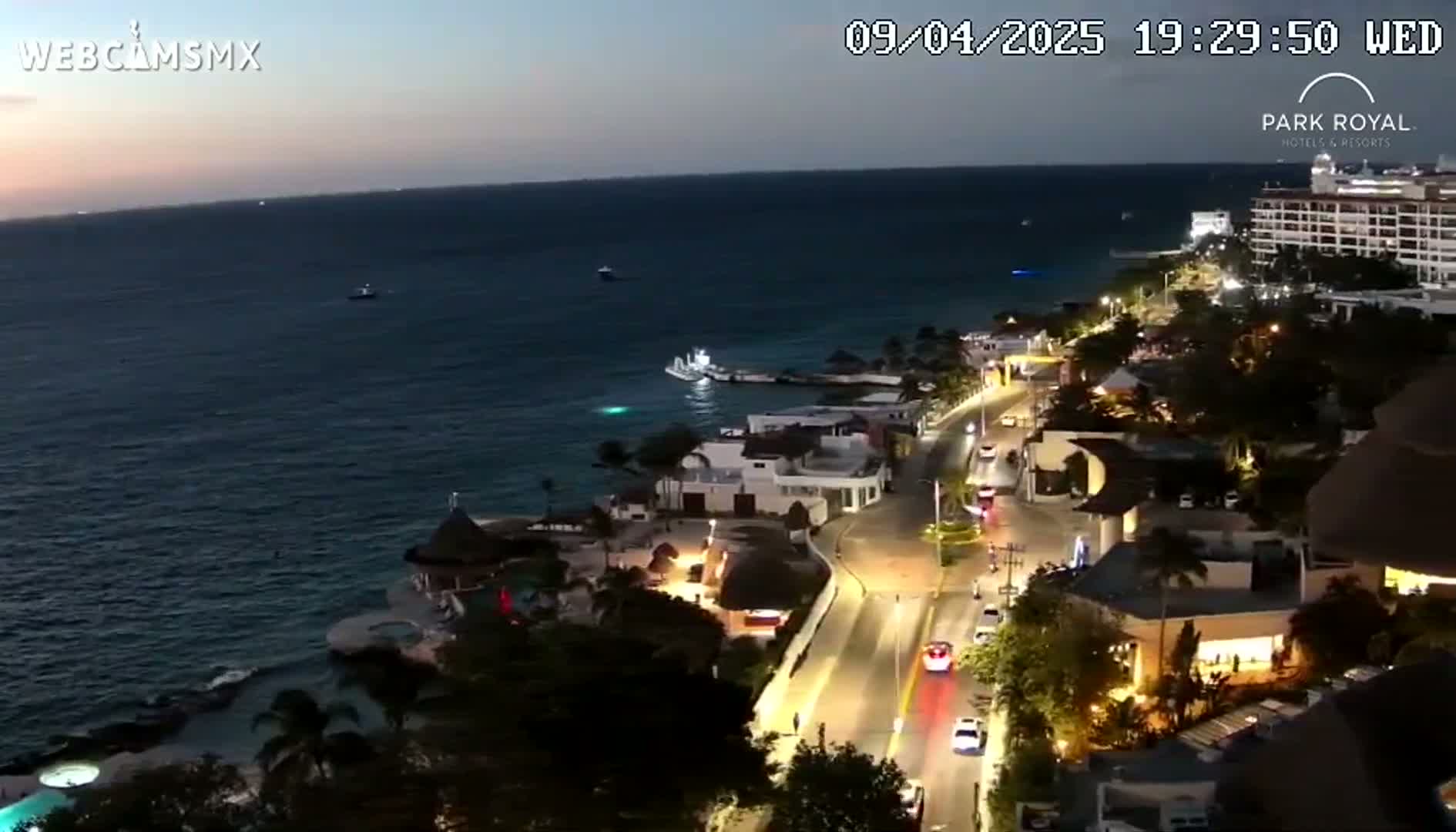 A nighttime coastal scene shows a road illuminated with streetlights winding alongside a dark body of water, with buildings and resorts lining the shore under a twilight sky.