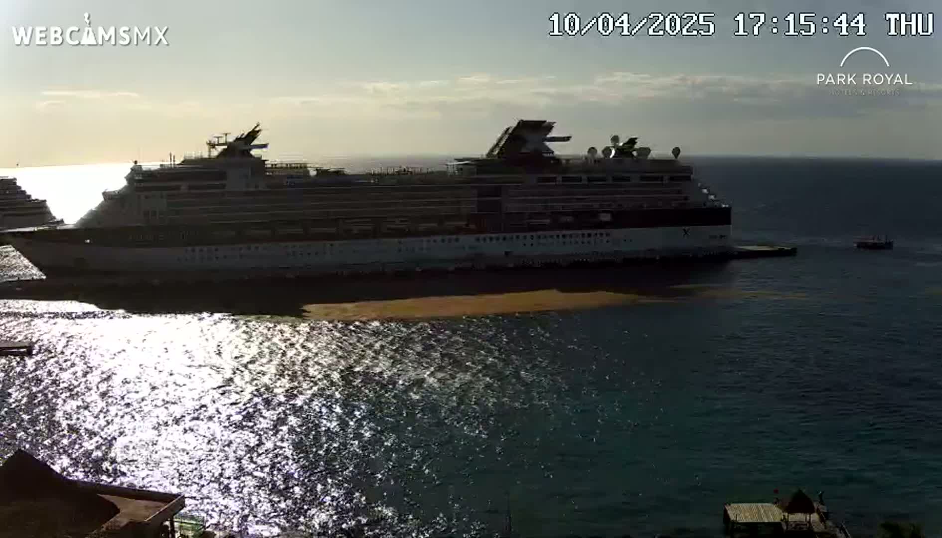 A large cruise ship is docked at a port, with another smaller ship visible in the distance, under a partly sunny sky.