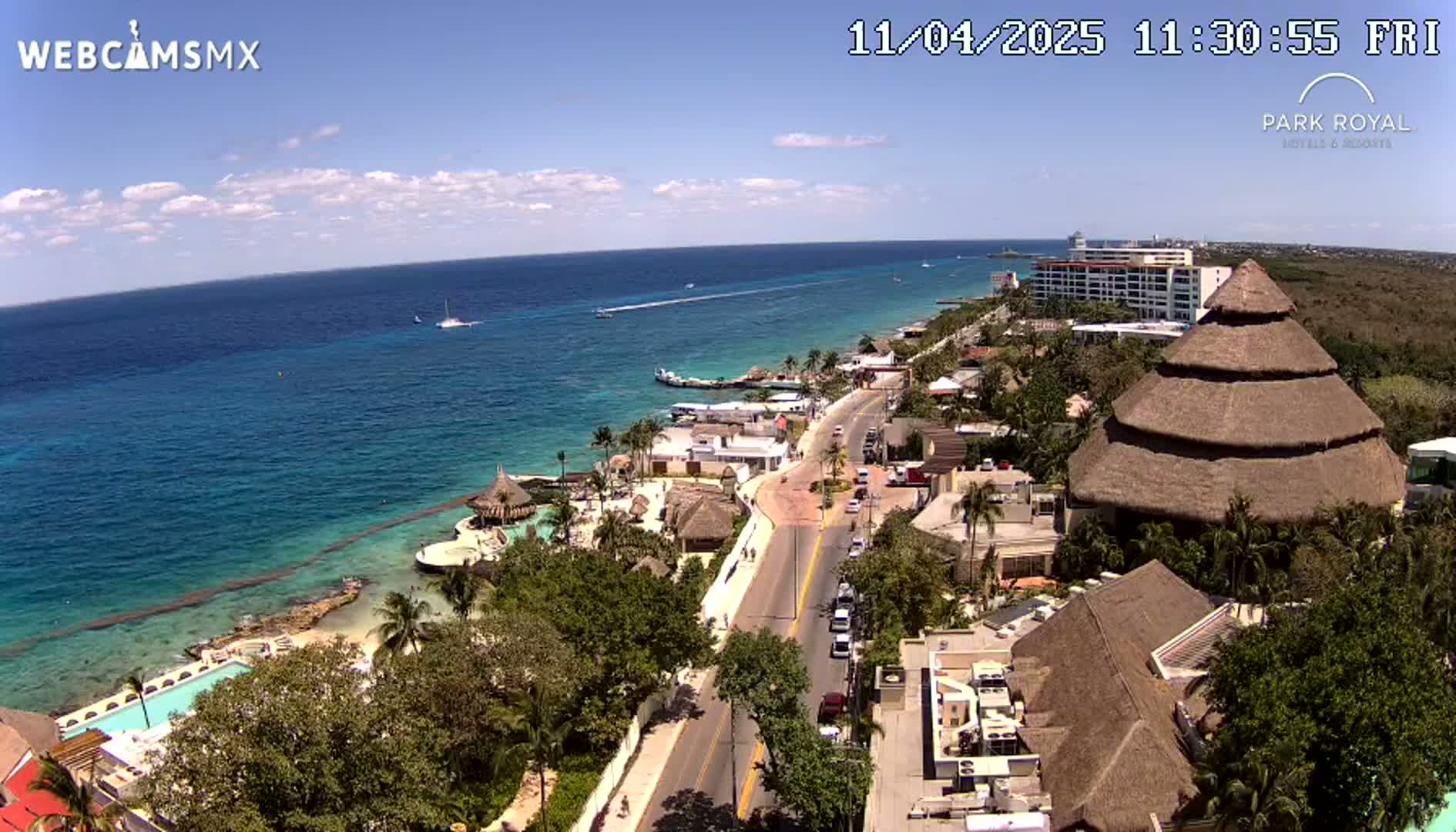 An aerial view of a tropical coastline on a sunny day shows a resort with thatched-roof buildings, a road, and turquoise ocean water with boats.