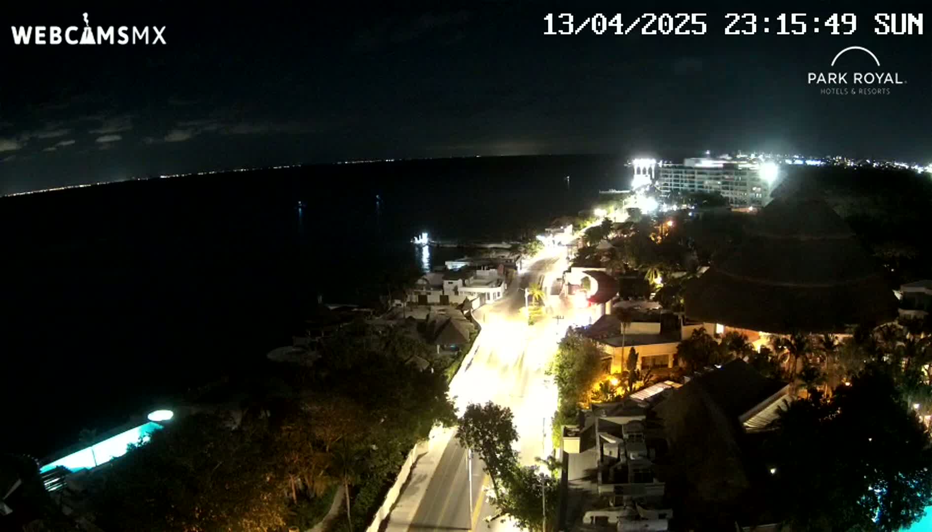 A nighttime coastal scene shows a brightly lit road winding through buildings near a dark body of water under a clear sky.