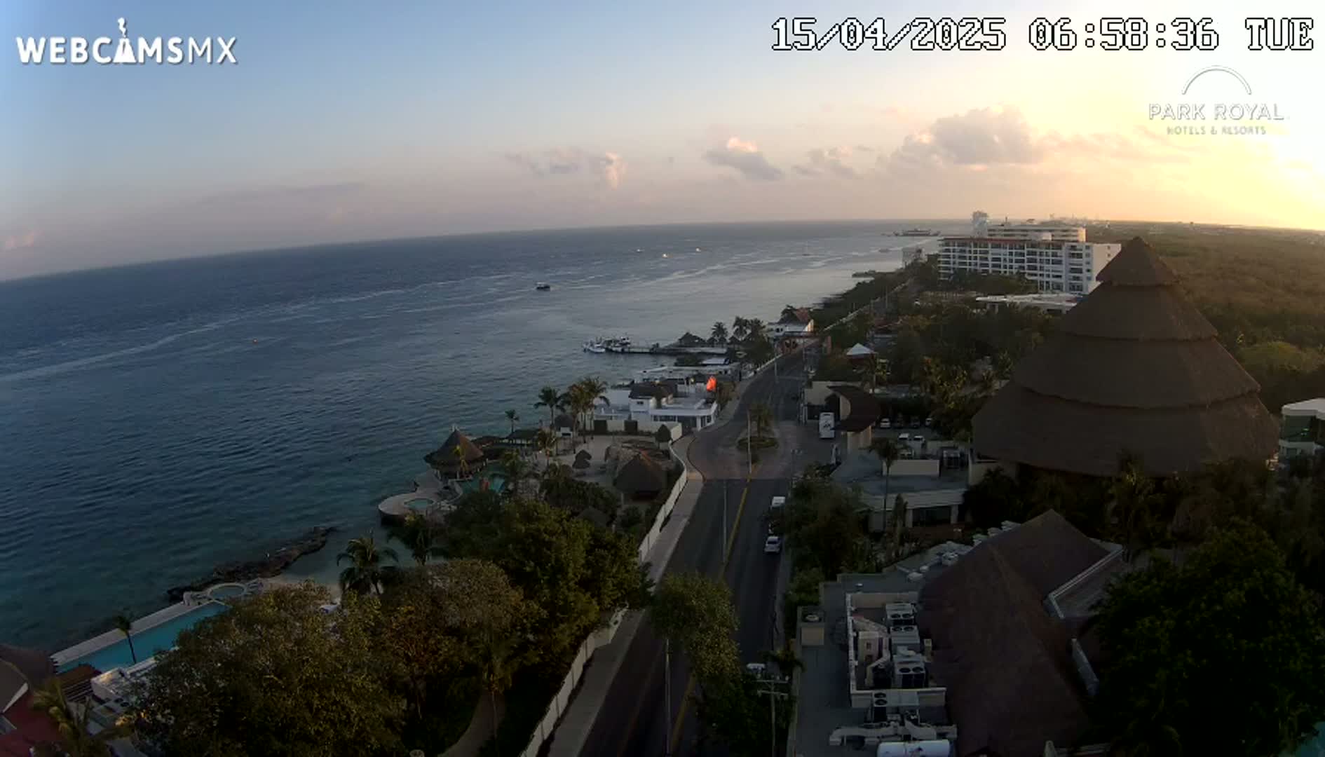 An aerial view at sunrise shows a coastal resort town with a calm ocean, a road, and several buildings, including a large thatched-roof structure.
