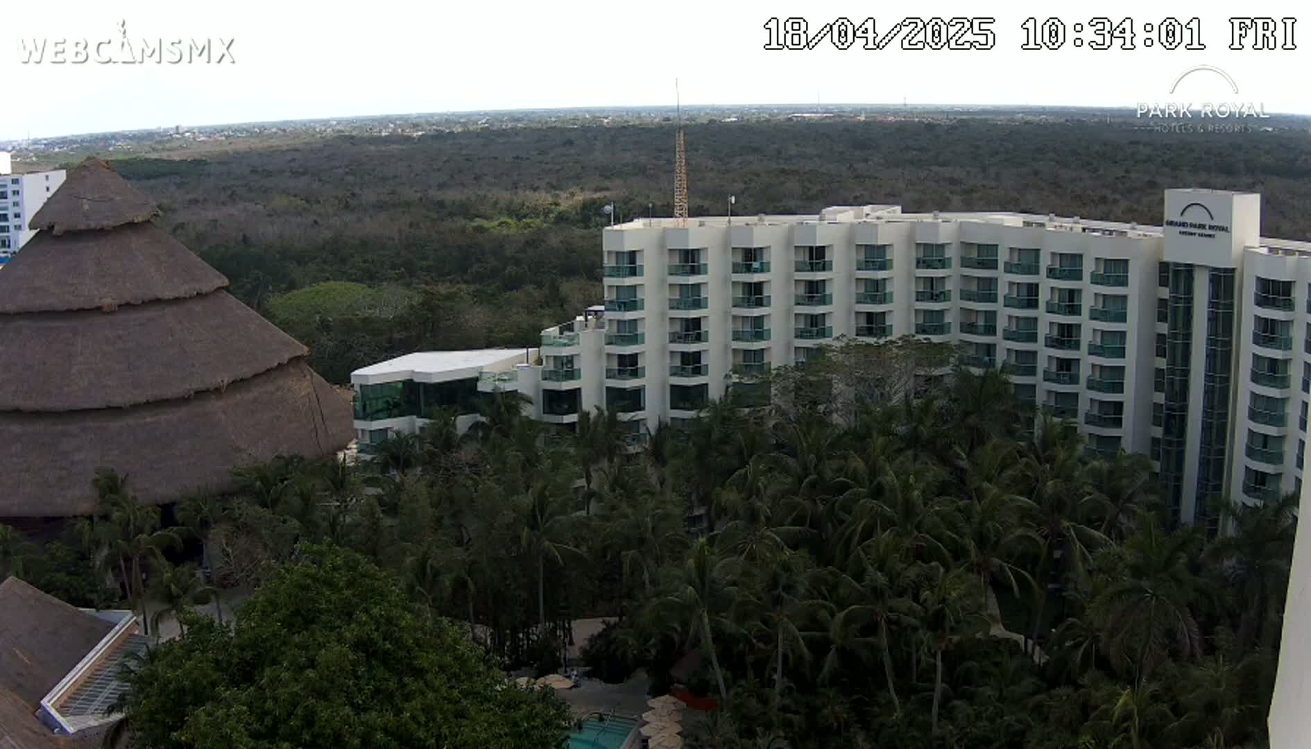 A large, multi-story hotel is situated amongst lush green tropical vegetation, with a thatched-roof structure visible in the foreground under a mostly clear sky.