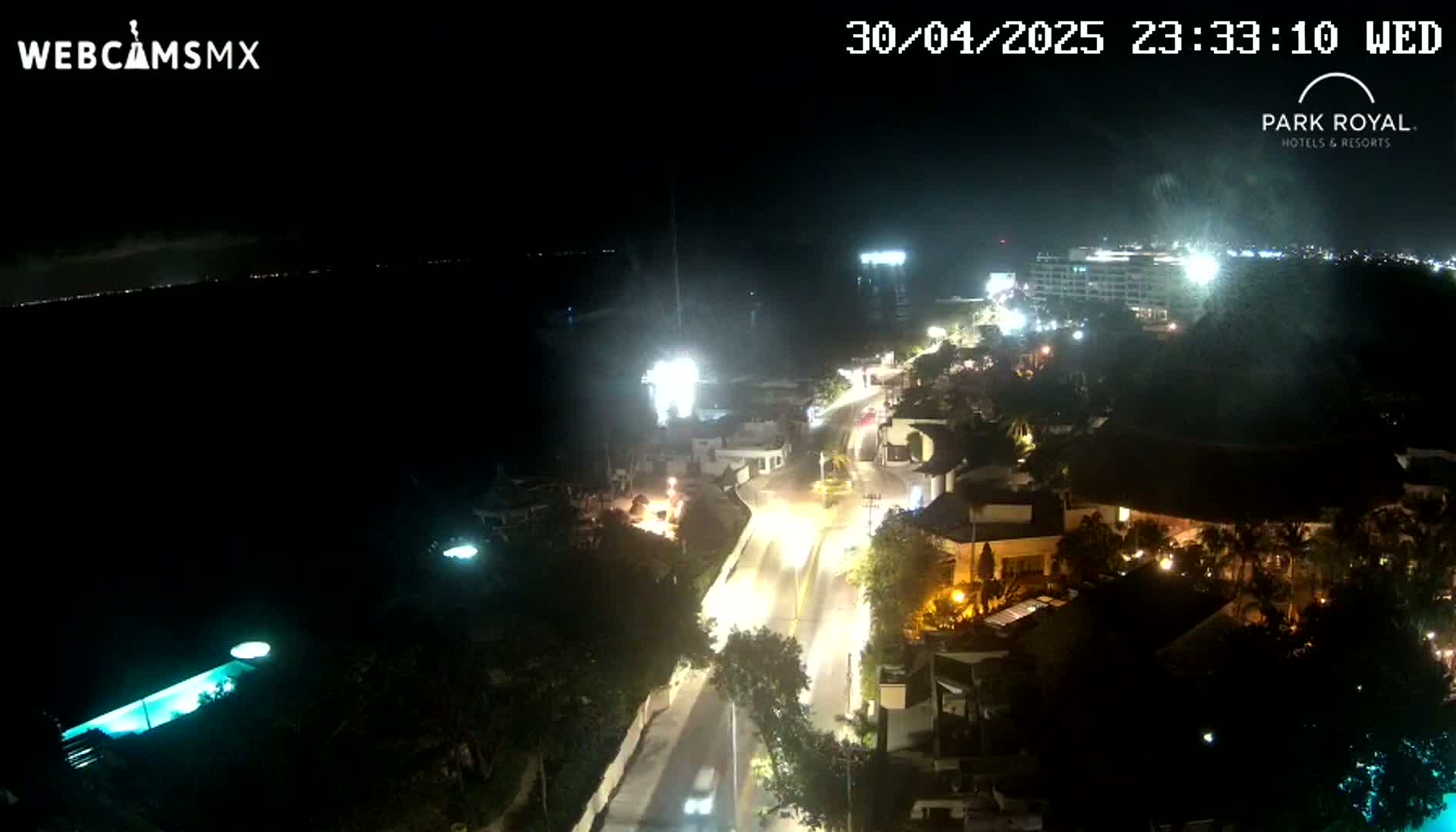 A nighttime aerial view shows a coastal road lined with buildings and lit by streetlights, with a dark ocean visible in the background.