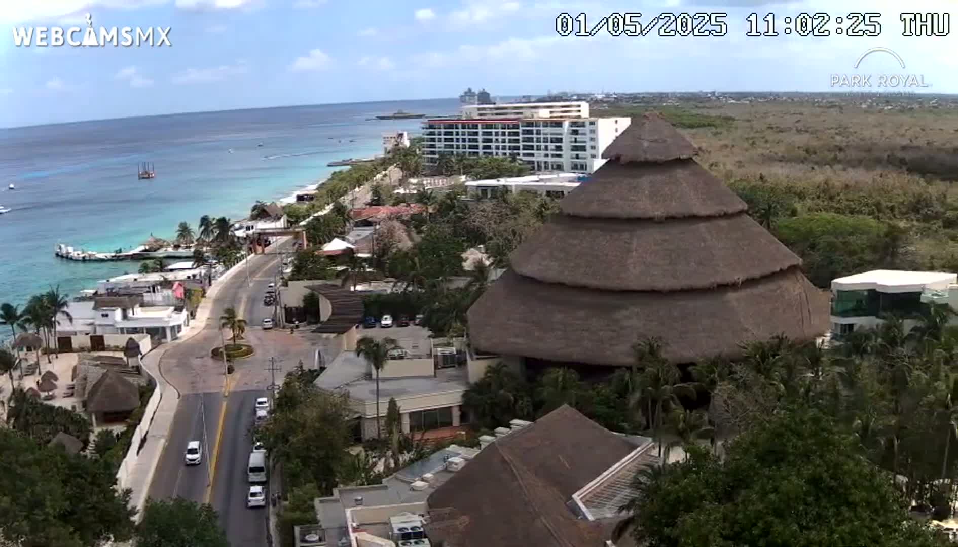 A coastal resort town on a sunny day, featuring a large thatched-roof building, hotels, and a road with cars, all overlooking a calm ocean.
