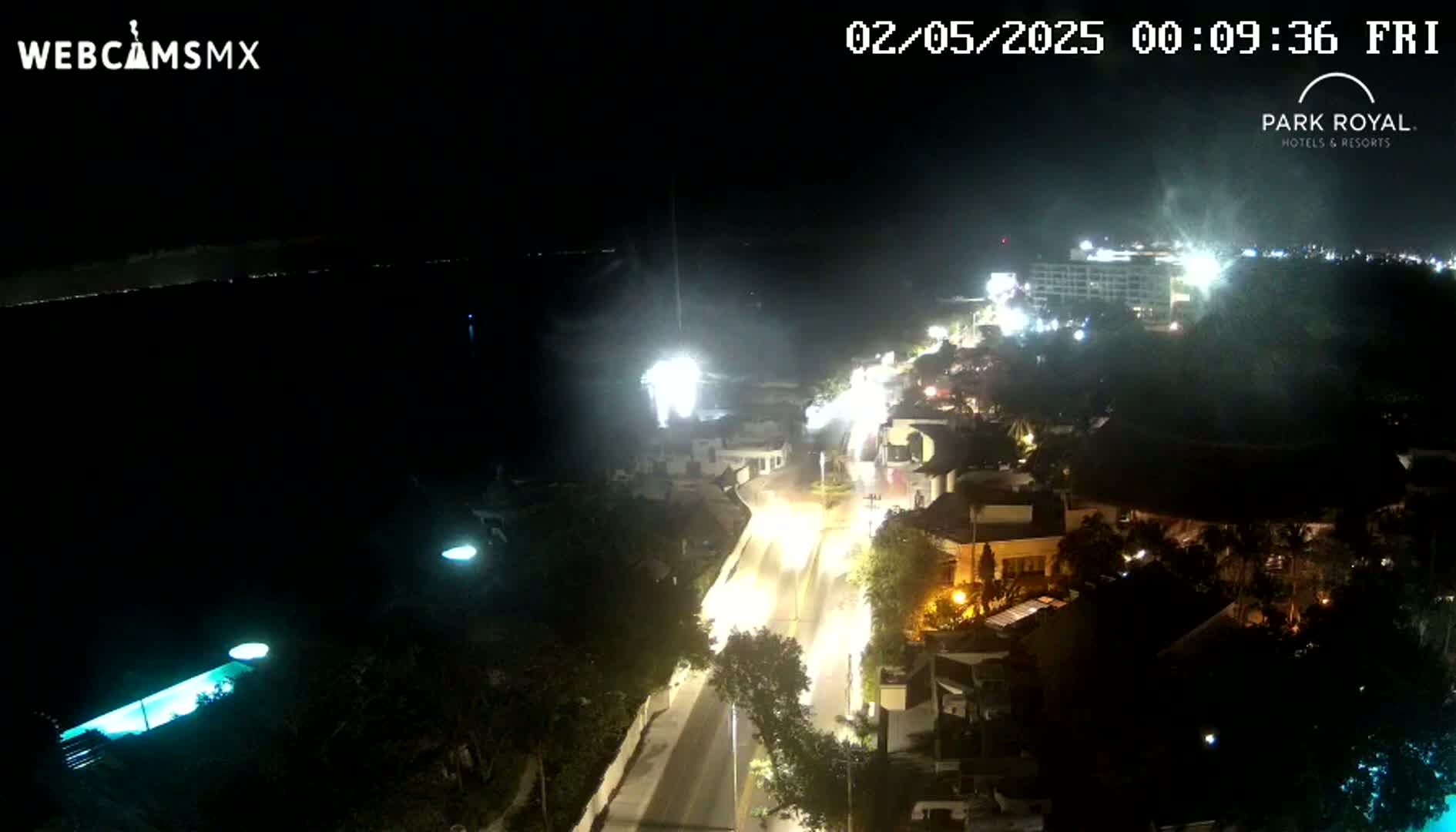 A nighttime aerial view shows a brightly lit coastal road and buildings, with a dark body of water visible in the background under a clear, dark sky.