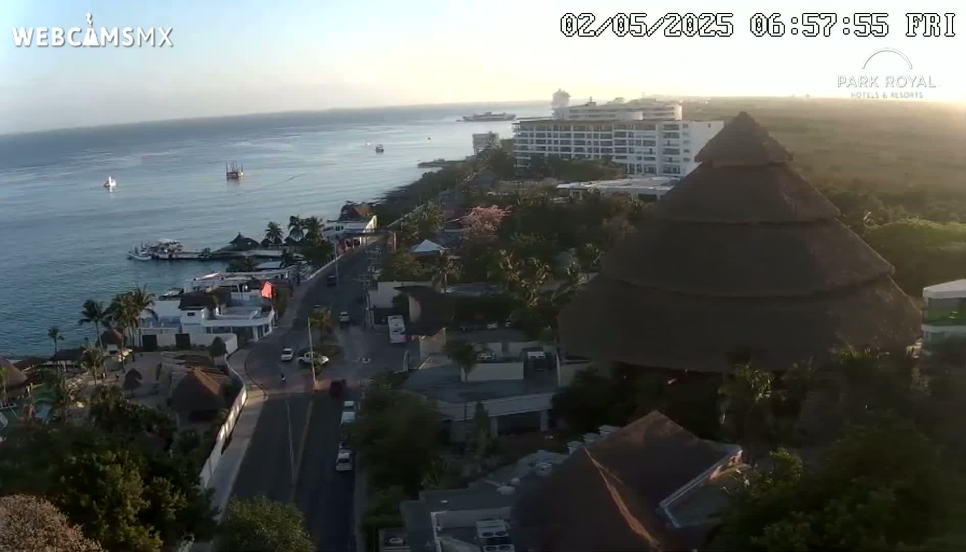 An aerial view shows a coastal town at sunrise with calm ocean waters, a large hotel, and a multi-tiered thatched roof structure.