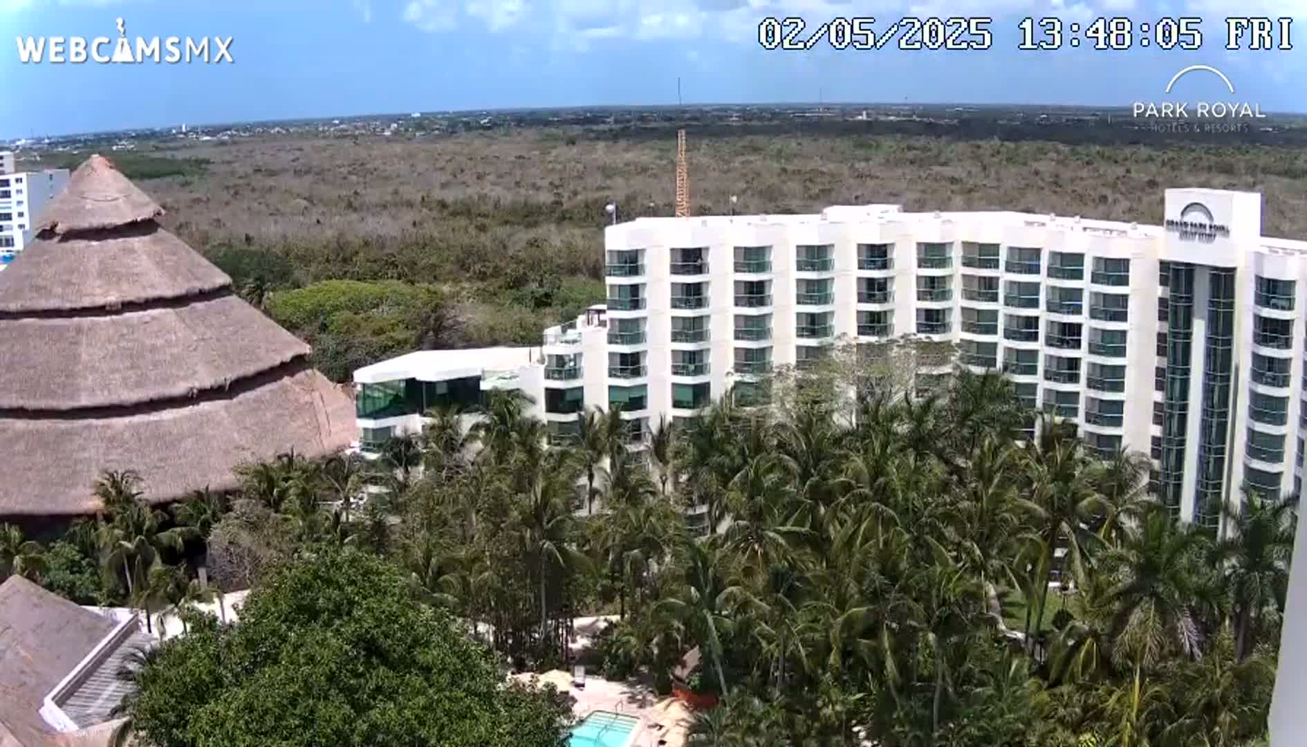 A large, multi-story hotel is surrounded by lush palm trees under a sunny sky, adjacent to a thatched-roof building.