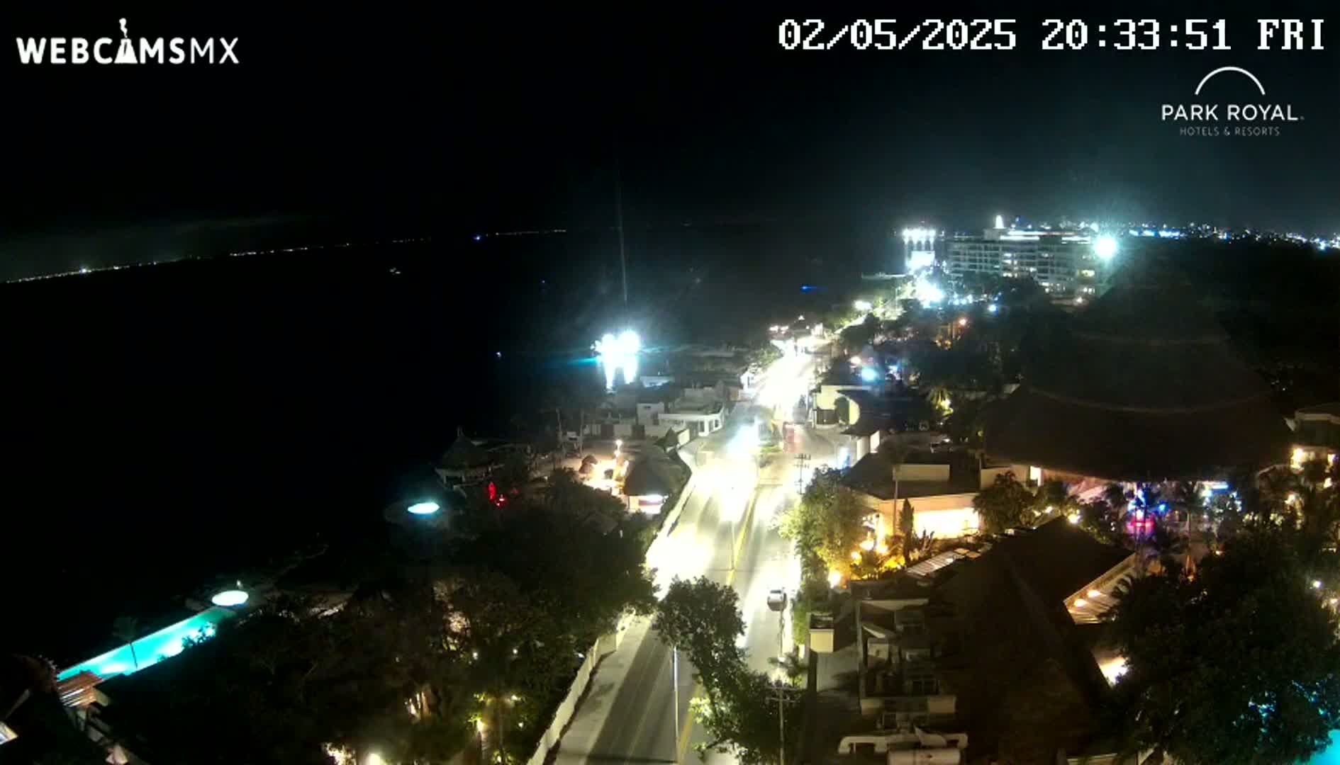 A brightly lit coastal town at night, seen from above, stretches along a road toward a dark ocean under a clear sky.