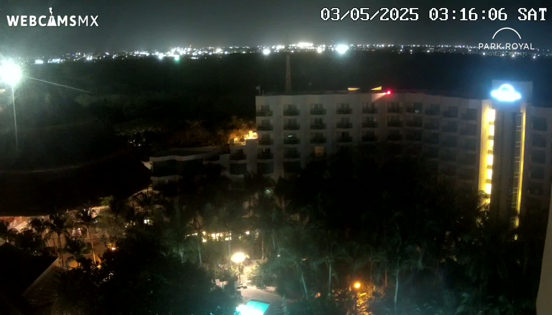 A brightly lit hotel complex at night, surrounded by palm trees and a swimming pool, with city lights visible in the distance under a clear, dark sky.