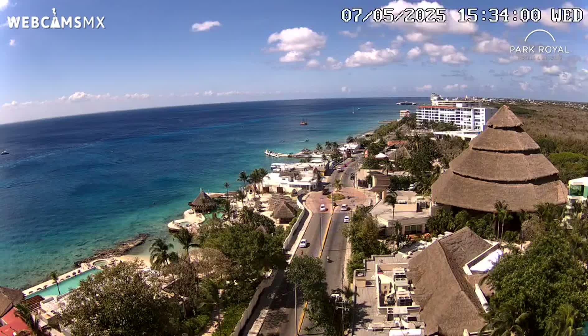 An aerial view shows a resort town on a sunny day, with a clear blue ocean in the background and a road winding through the buildings toward the shore.