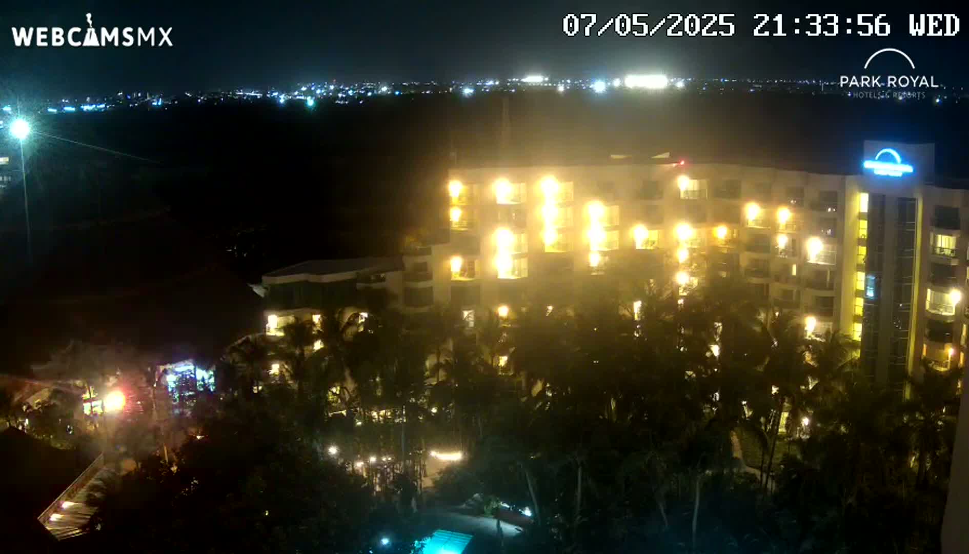 A brightly lit multi-story hotel surrounded by palm trees at night, with a city skyline visible in the distance under a clear sky.