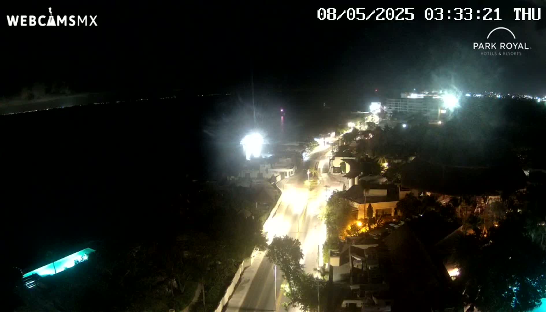 A nighttime, overhead view shows a coastal road with vehicle lights winding through buildings and vegetation towards a dark ocean under a partly cloudy sky.