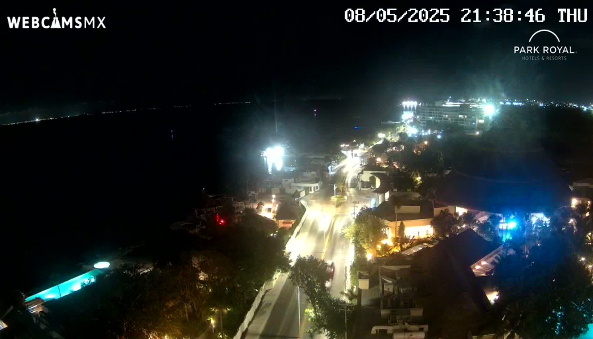 A nighttime aerial view of a coastal town shows a brightly lit road leading to a large resort and oceanfront buildings, with a dark body of water visible in the background under a clear sky.