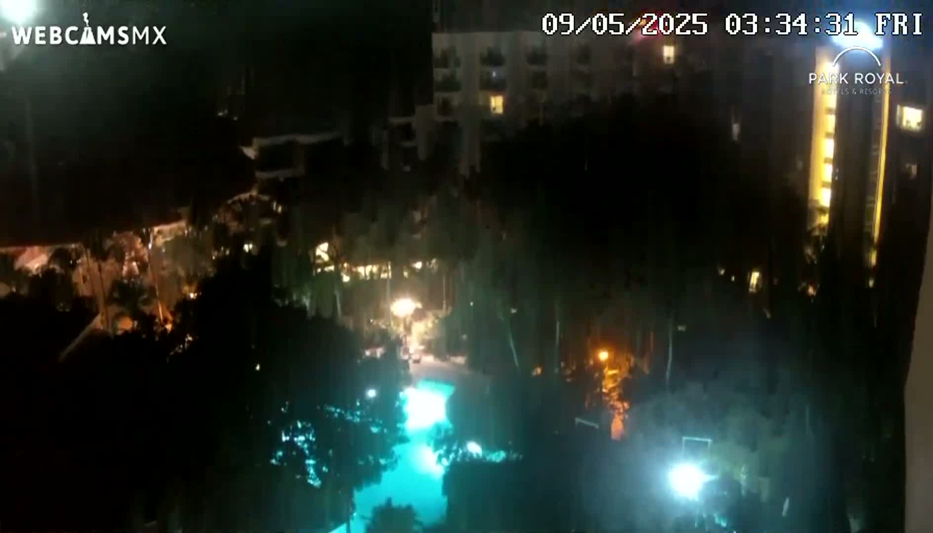 A nighttime aerial view shows a resort pool lit in teal, surrounded by dark foliage and illuminated buildings.