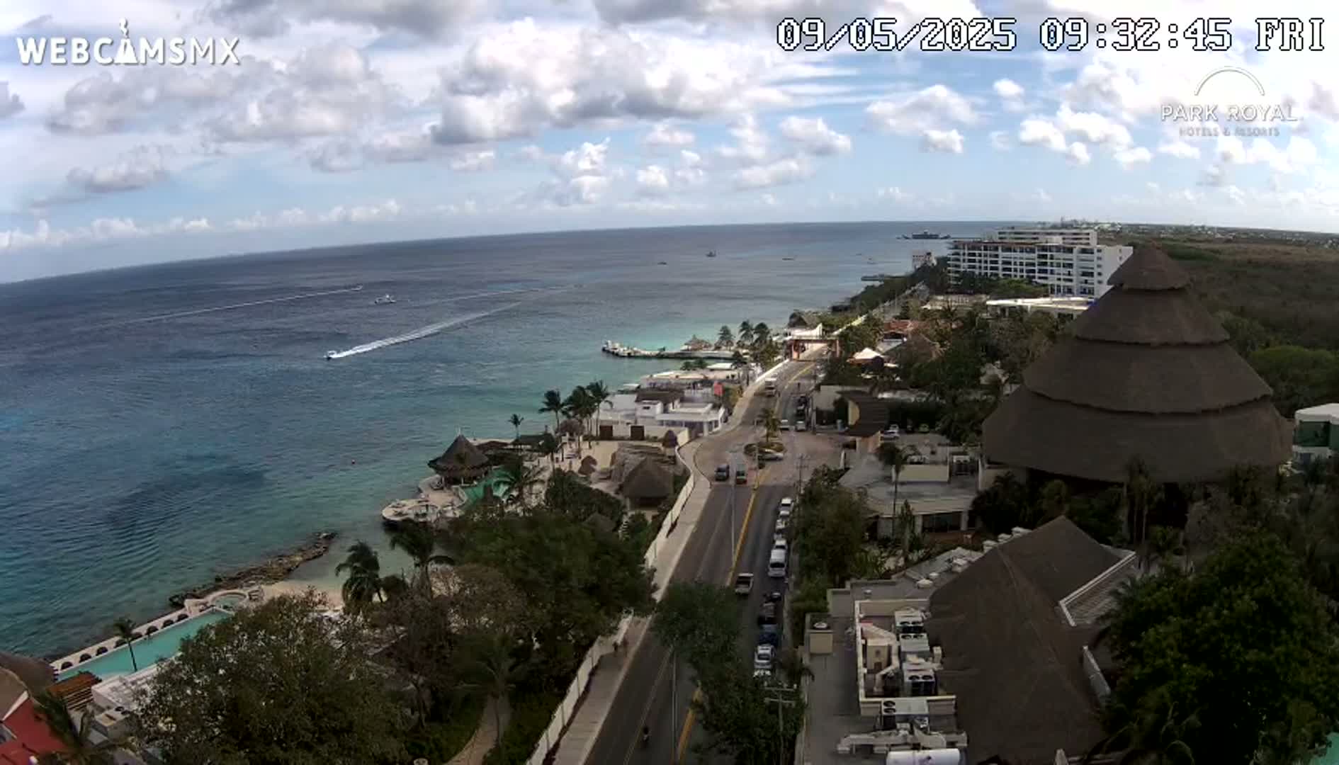 An aerial view shows a coastal resort town on a partly cloudy day, with boats visible in the ocean and a large thatched-roof structure prominent in the foreground.