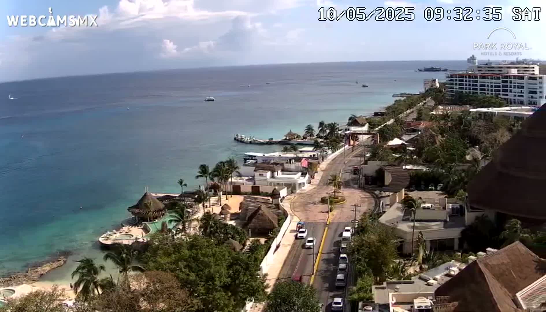 A mostly sunny day shows a coastal town with turquoise water, palm trees, buildings, and a road with cars, viewed from an elevated perspective.