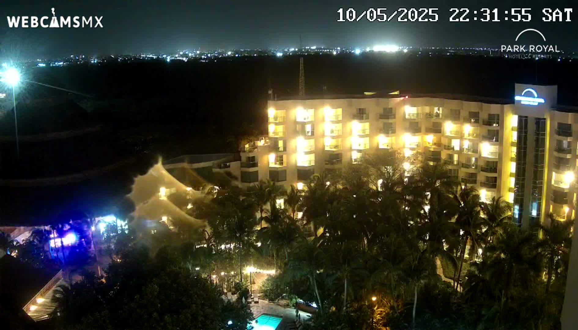 A brightly lit hotel at night is surrounded by palm trees, with a large spray of water visible near the foreground under a clear night sky.