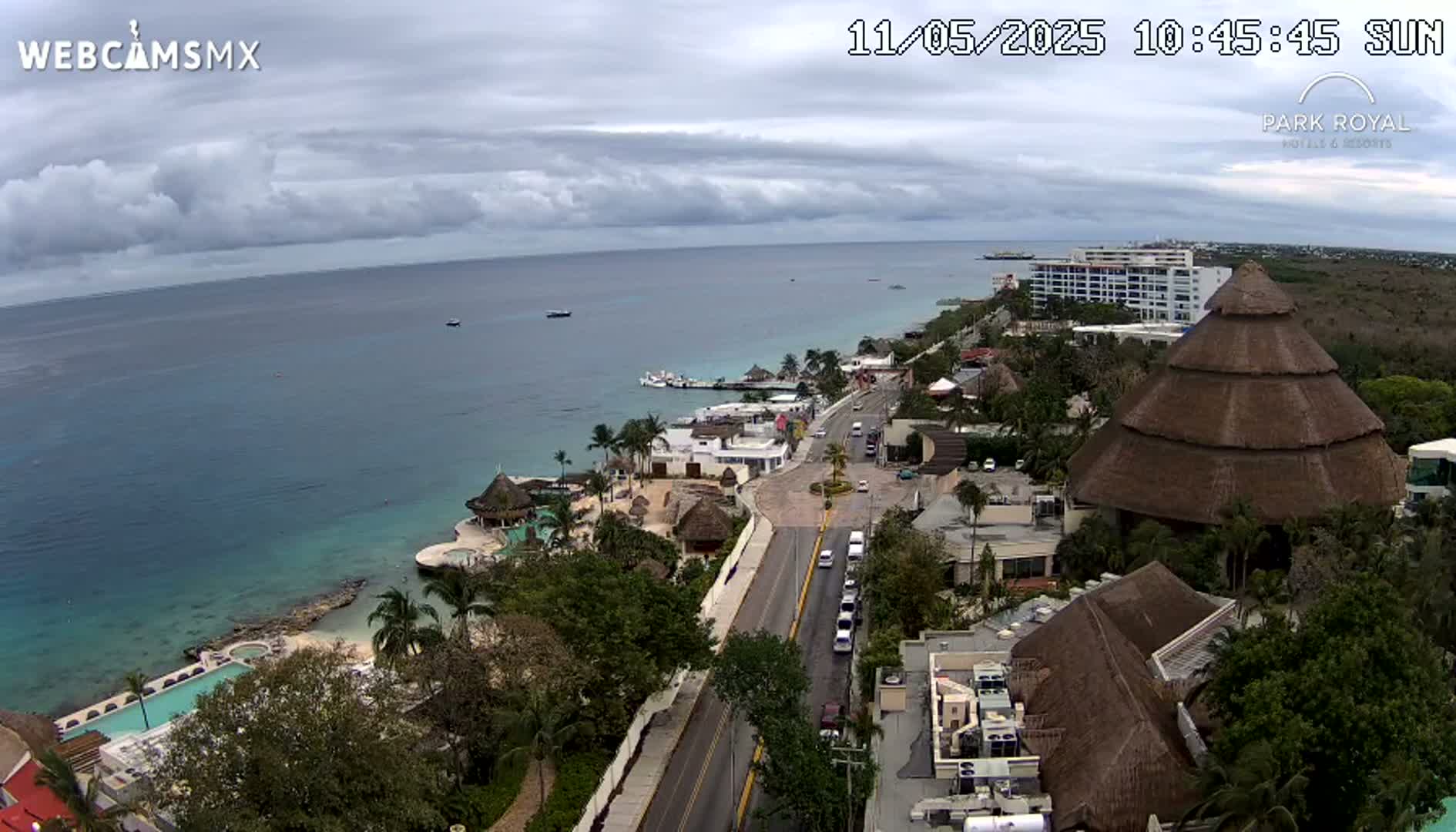 An aerial view shows a coastal resort town on a partly cloudy day, with a turquoise ocean, a road leading to the beach, and several buildings including a large thatched-roof structure.