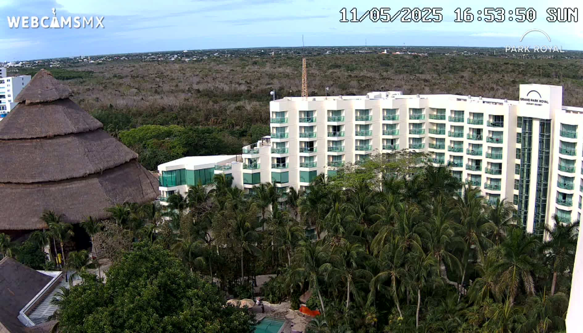 A large resort hotel complex with a thatched-roof building is nestled among lush tropical vegetation under a partly cloudy sky.