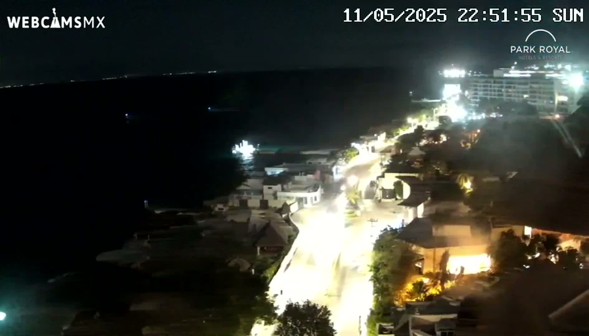 A nighttime, overhead view shows a brightly lit coastal road winding through a town beside a dark ocean.