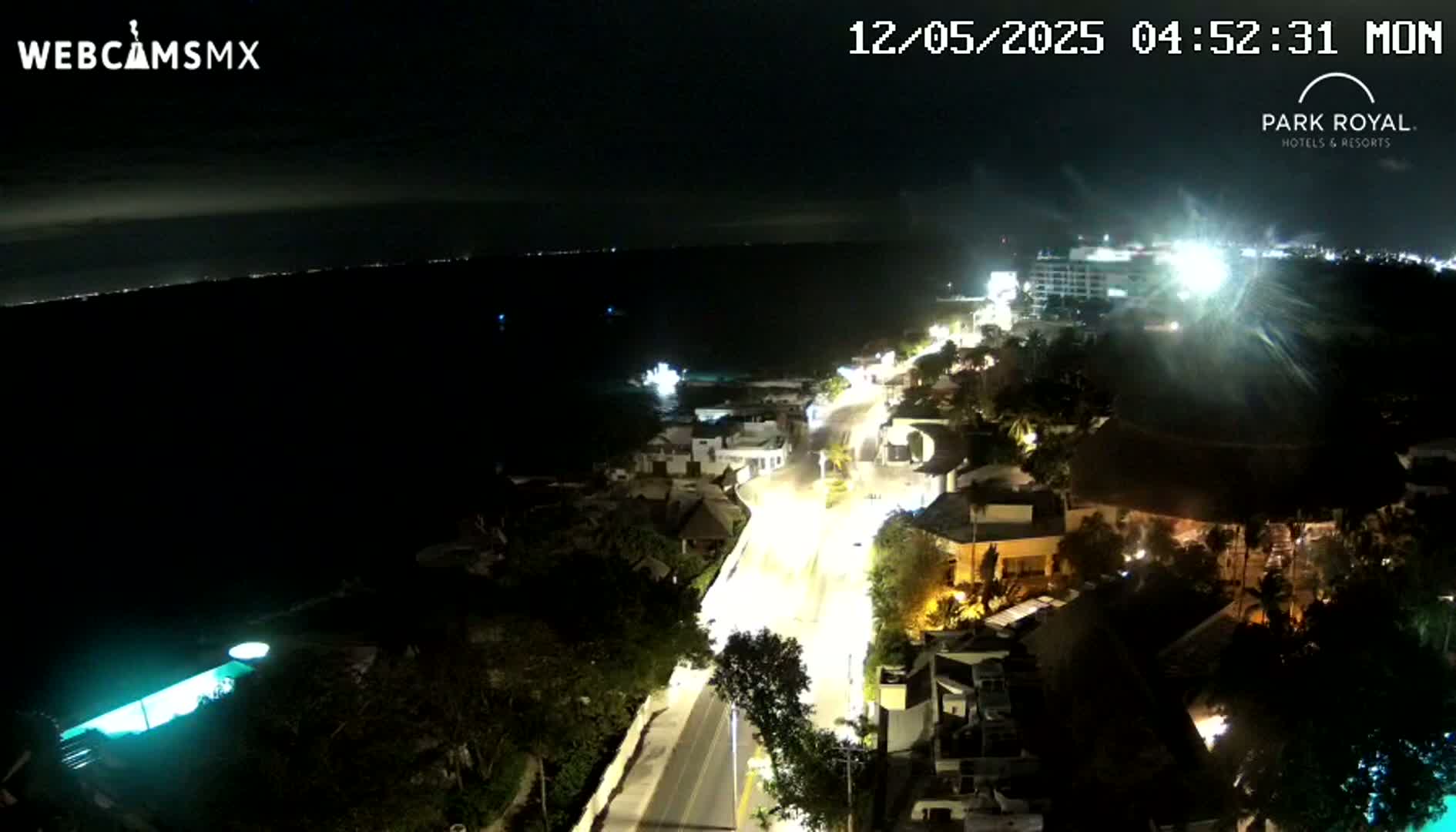 A nighttime, high-angle view shows a coastal road illuminated by streetlights, leading to a dark ocean under a mostly clear sky.