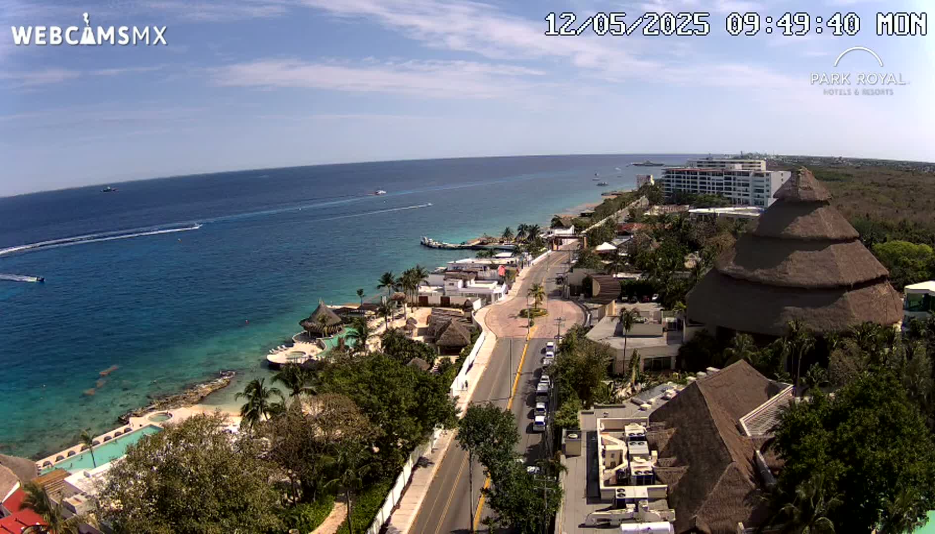 An aerial view shows a coastal resort town on a sunny day, with a turquoise ocean, a road lined with buildings, and a large thatched-roof structure.