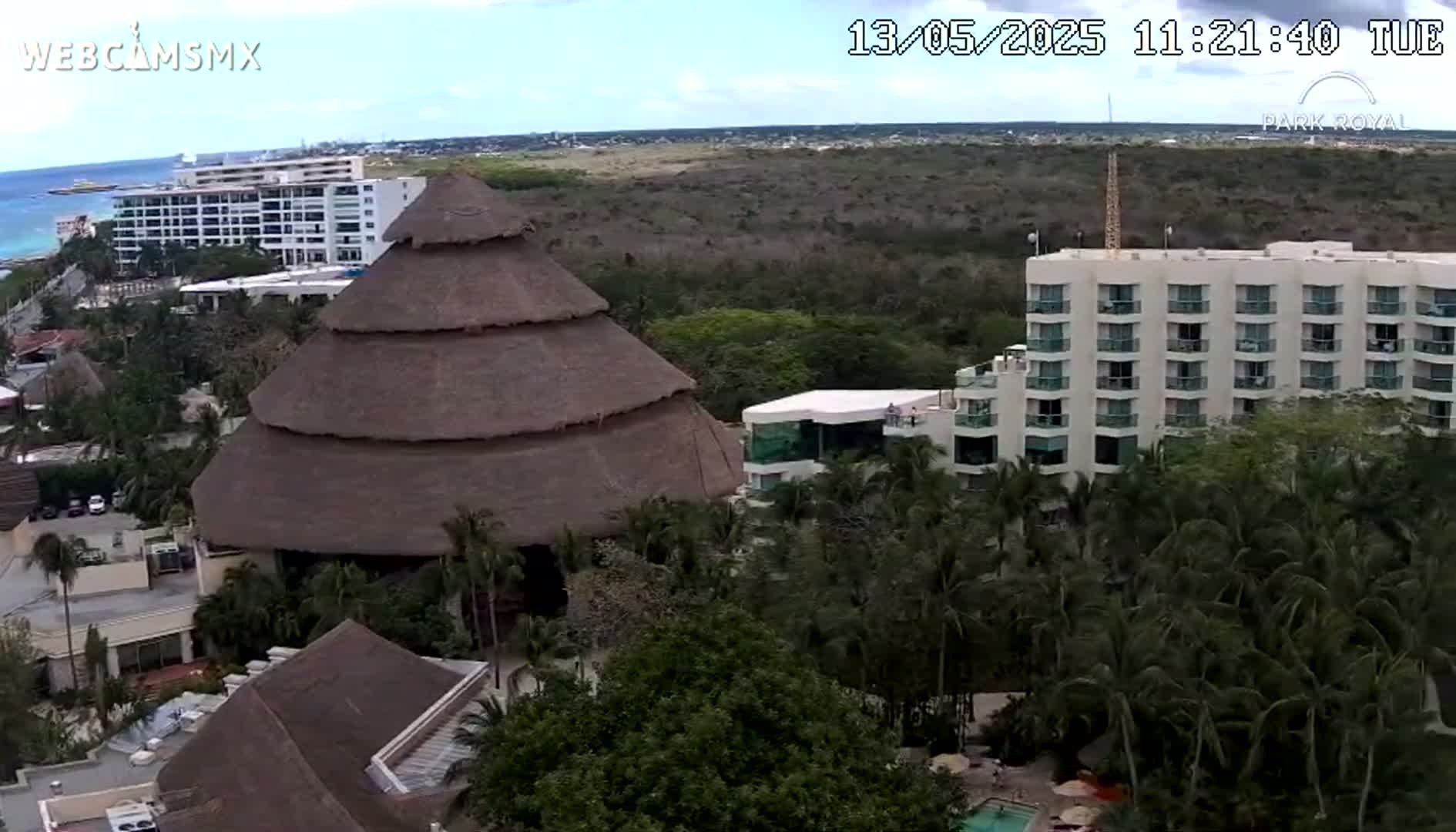 An aerial view shows a resort with several buildings, including a large thatched-roof structure, under partly cloudy skies.