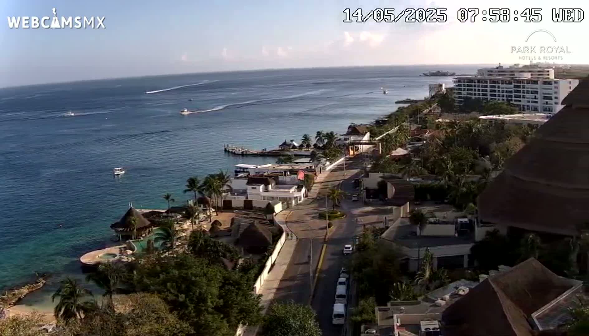 An elevated view shows a coastal road winding past tropical vegetation, resorts, and a calm ocean with several small boats under a clear, sunny sky.