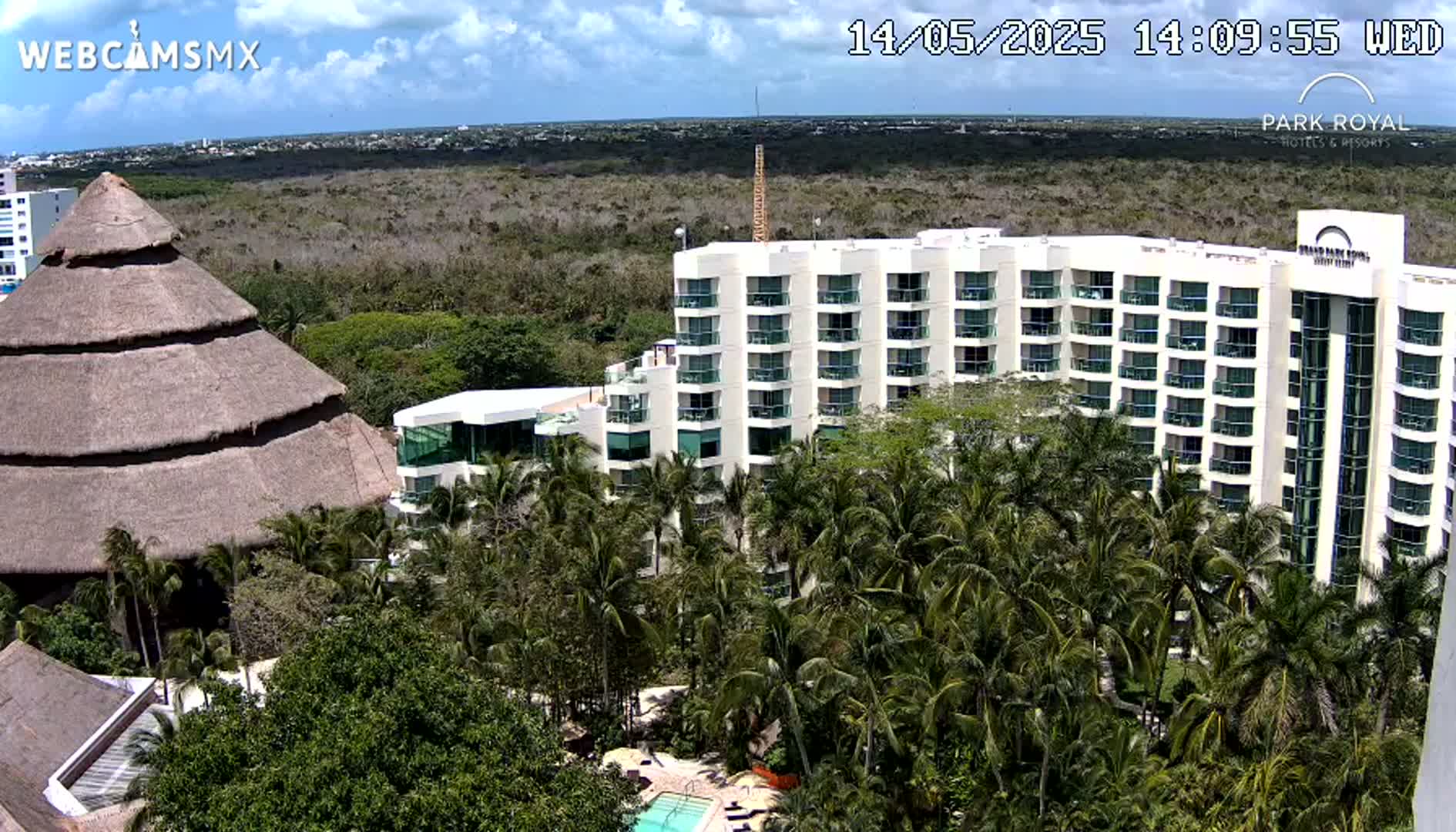 A large hotel complex with a thatched-roof building sits amongst lush tropical vegetation under partly cloudy skies.