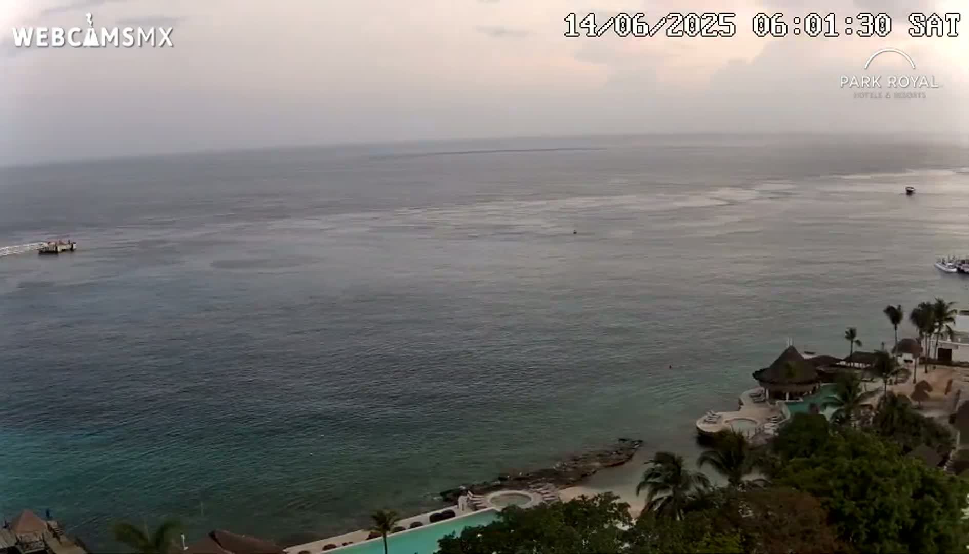 A calm, overcast ocean is visible from an elevated vantage point, overlooking a resort with a pool and palm trees on a rocky shoreline.