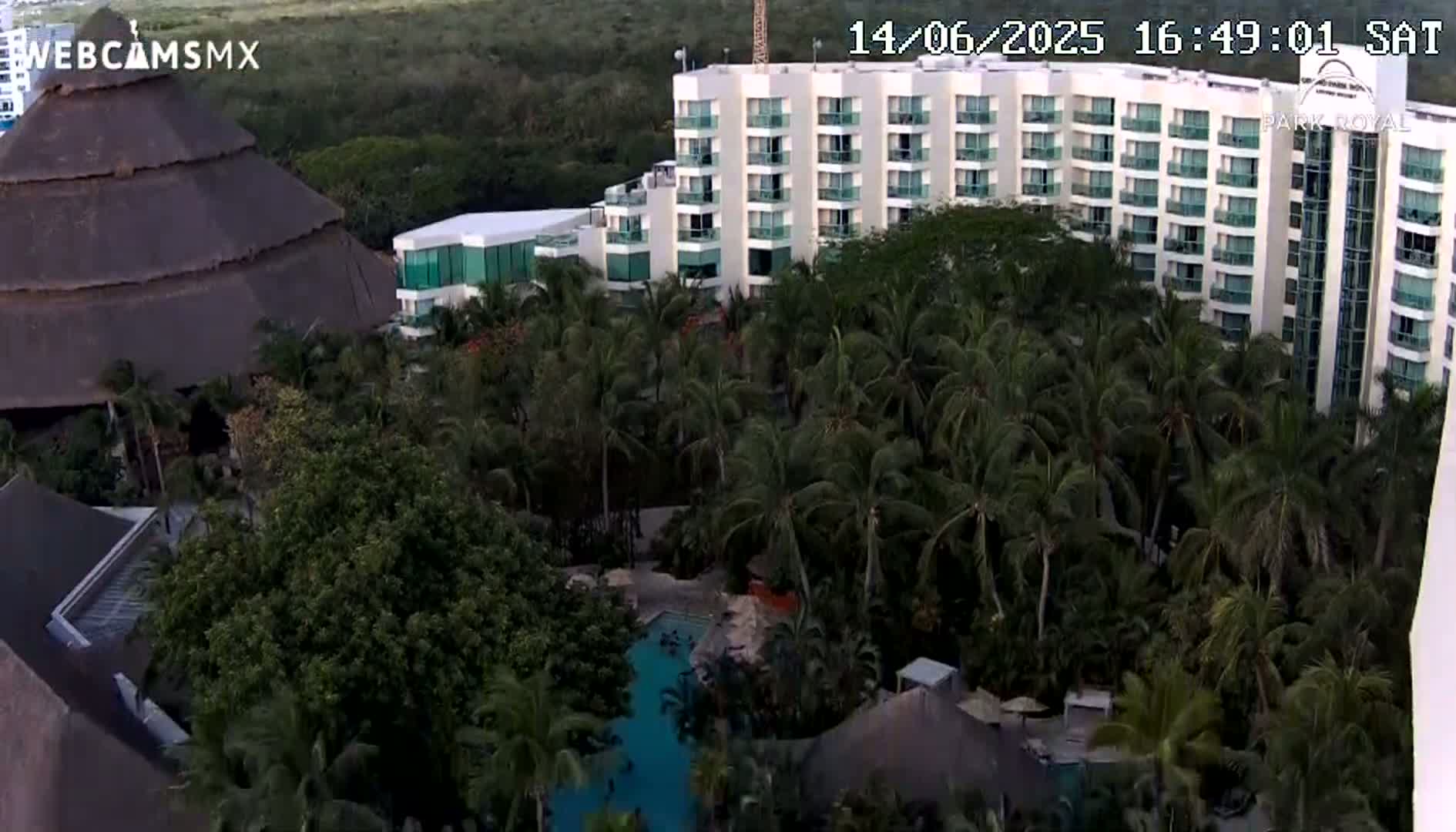 A calm, overcast ocean is visible from an elevated vantage point, overlooking a resort with a pool and palm trees on a rocky shoreline.