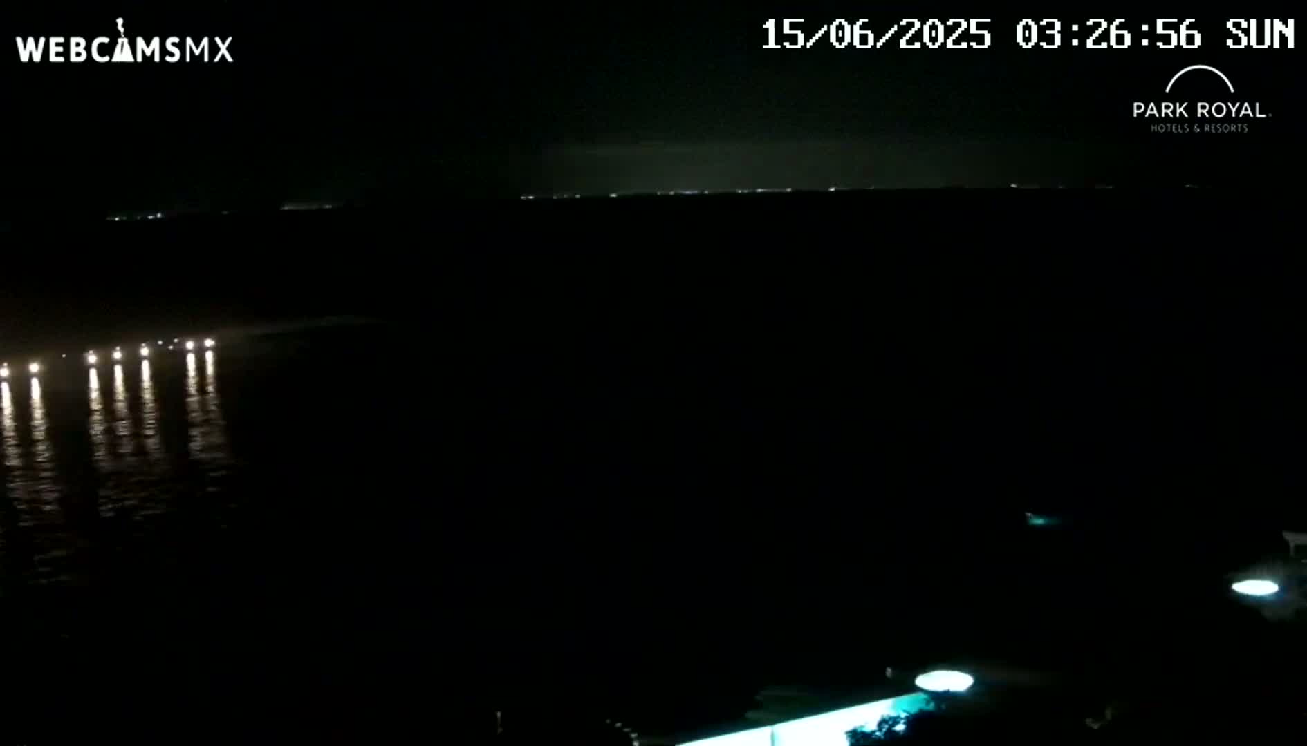 A nighttime view of a dark body of water with a line of lights reflecting on its surface, and a faintly visible shoreline in the distance.
