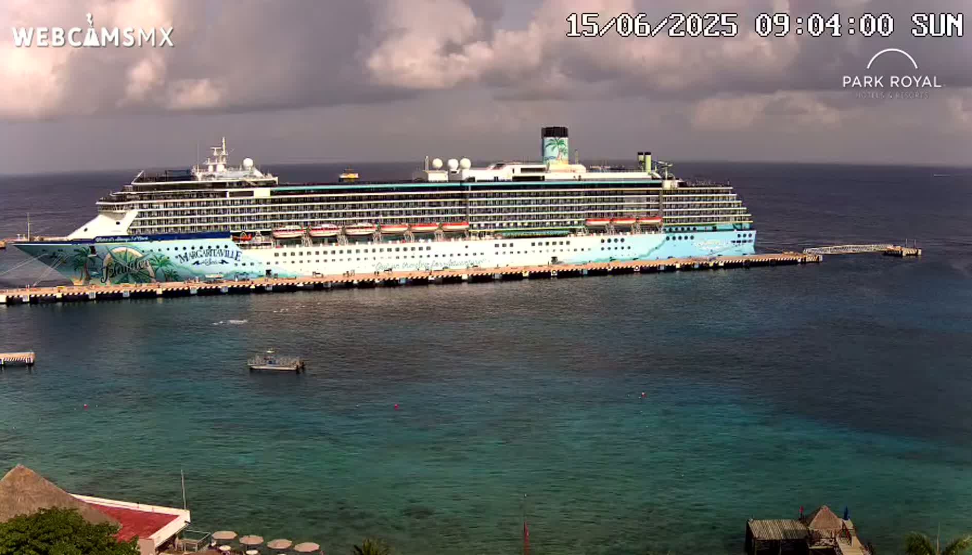 A large cruise ship is docked at a pier on a partly cloudy day, with clear, turquoise water surrounding it.