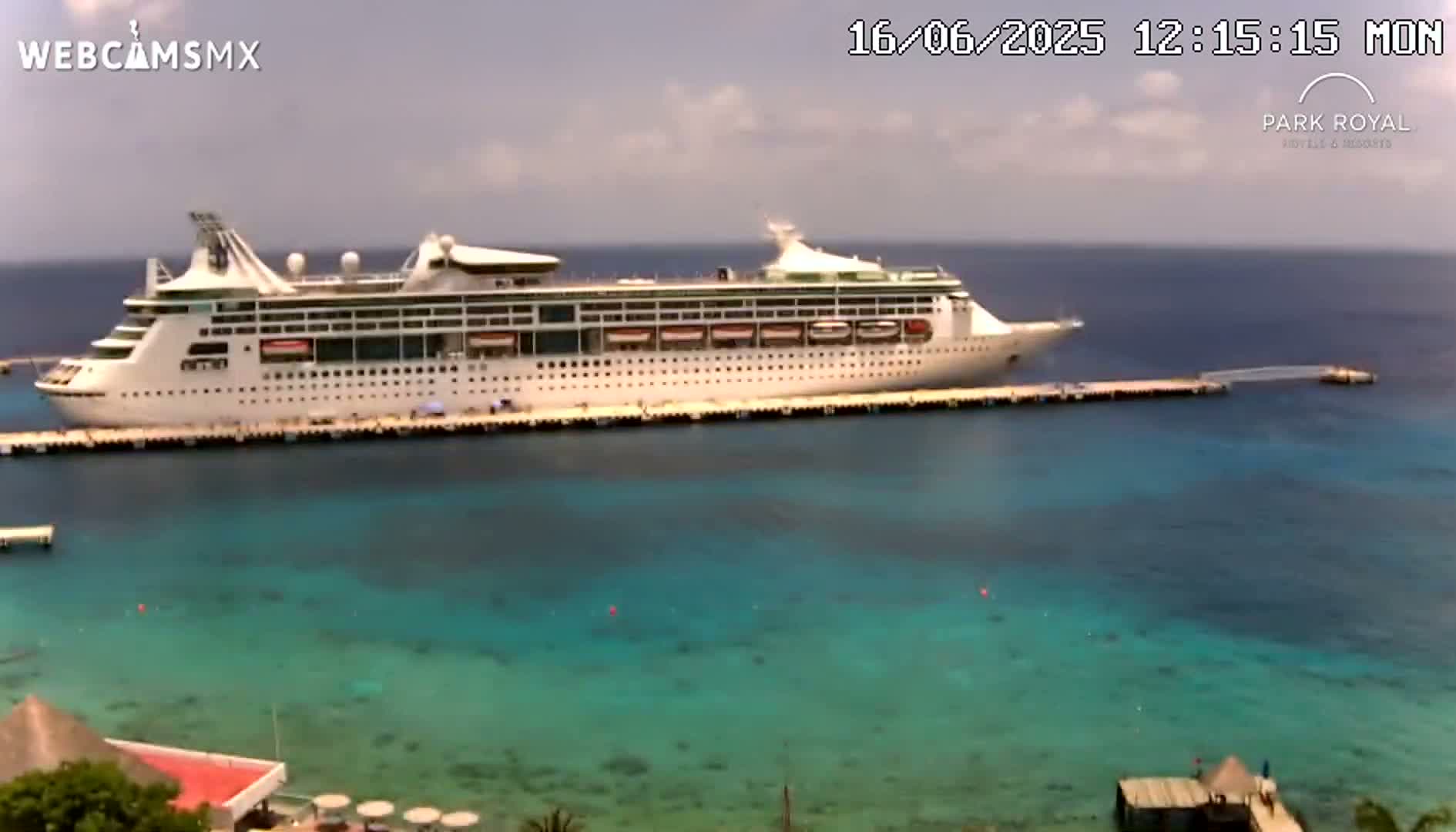 A large cruise ship is docked at a pier in calm, clear turquoise water under a mostly cloudy sky.