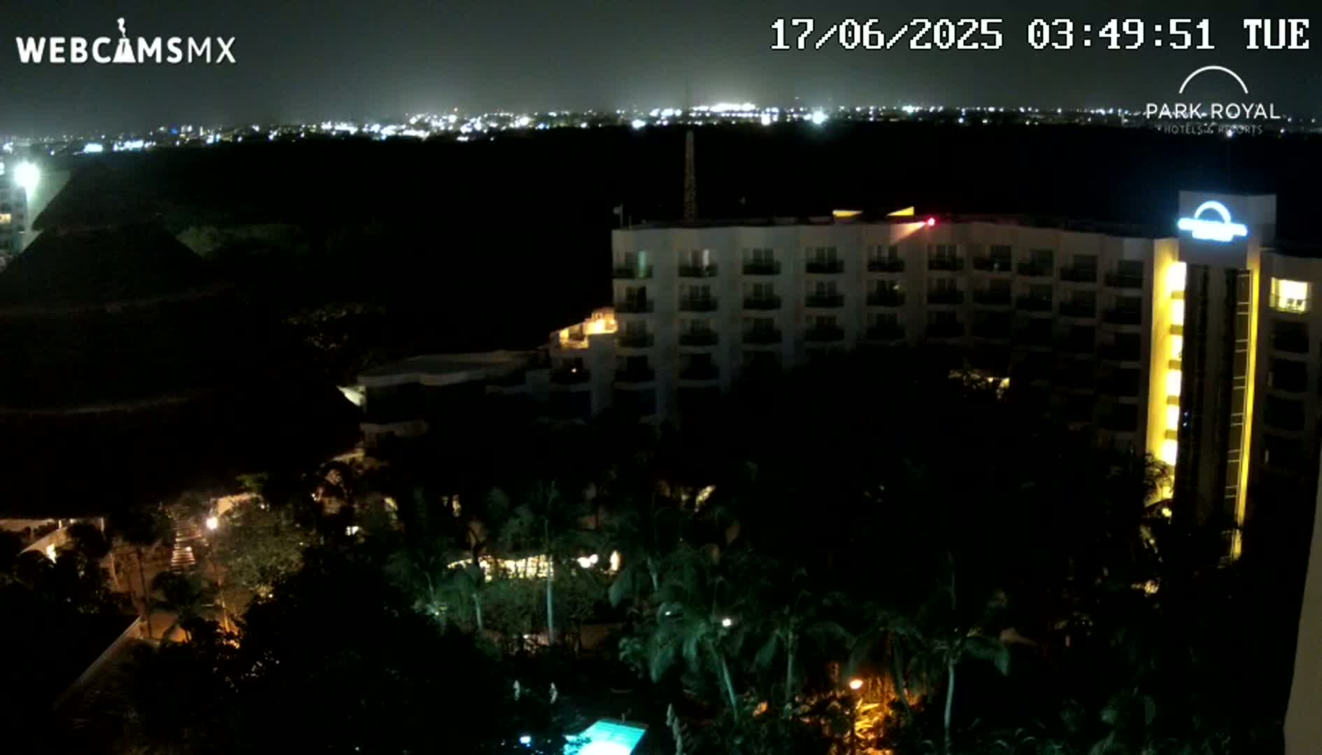 A large, multi-story hotel building is illuminated at night, surrounded by dark tropical vegetation, with a distant city skyline visible in the background under a clear night sky.