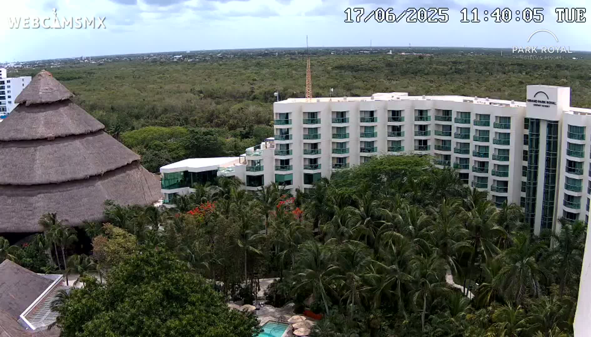 A large resort hotel complex with a thatched-roof building is nestled among lush tropical vegetation under partly cloudy skies.
