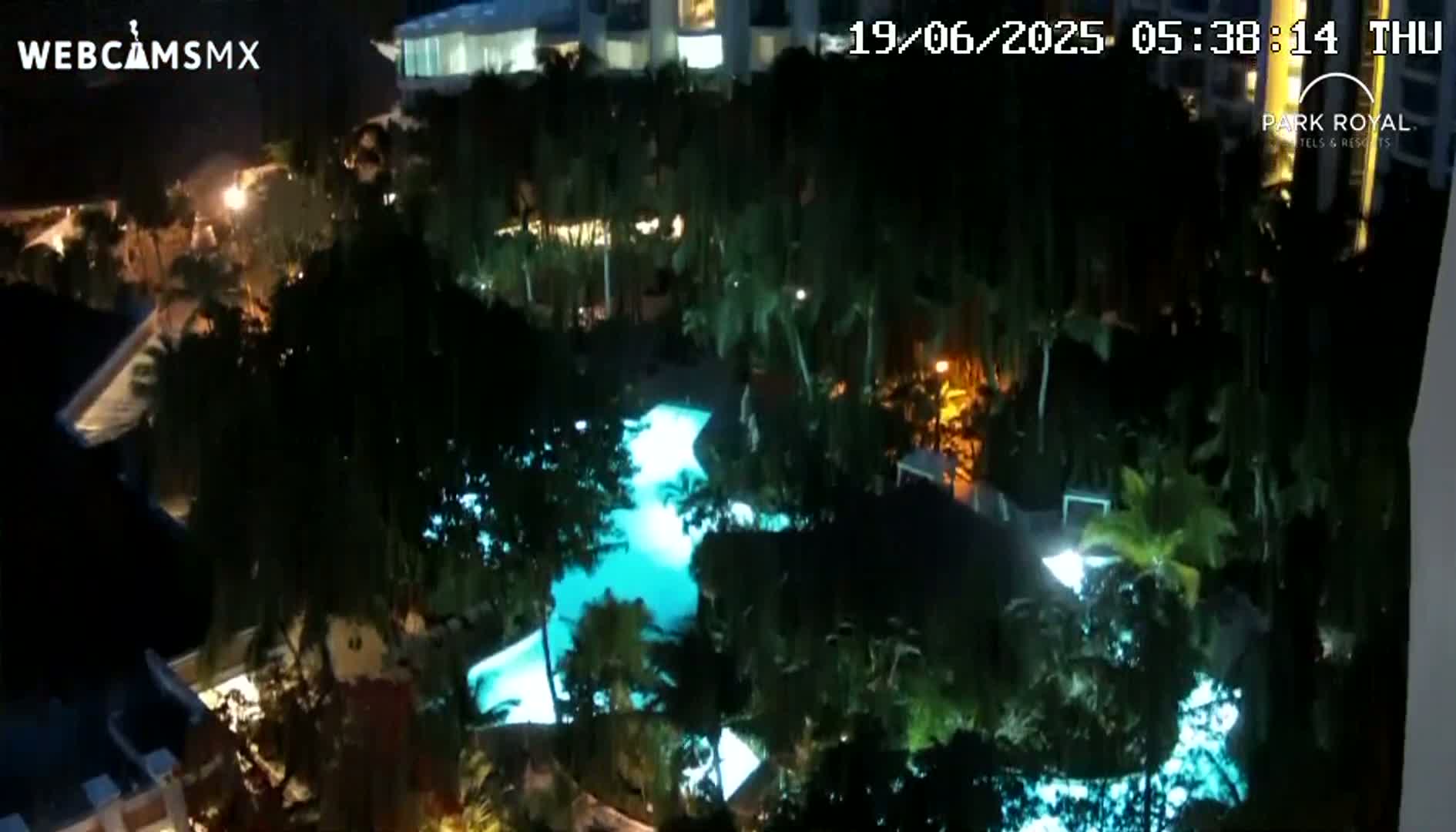A nighttime aerial view shows a resort pool lit up in turquoise, surrounded by lush, dark green tropical foliage.