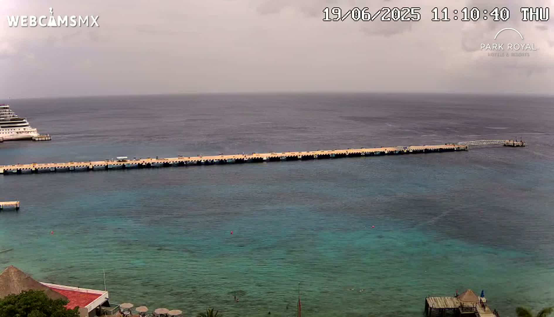 A long pier extends into a calm, teal ocean where a cruise ship is docked under an overcast sky.