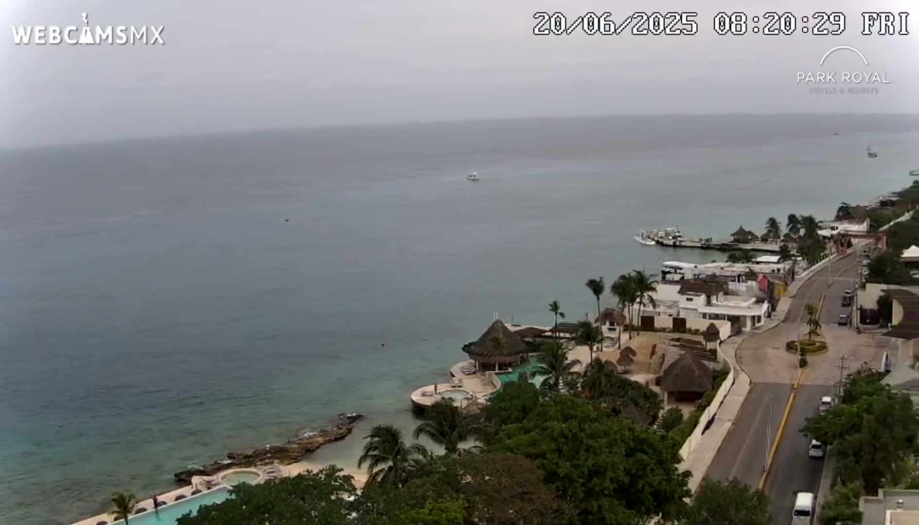 An overcast day shows a coastal resort town with a calm sea, a road, and tropical vegetation.