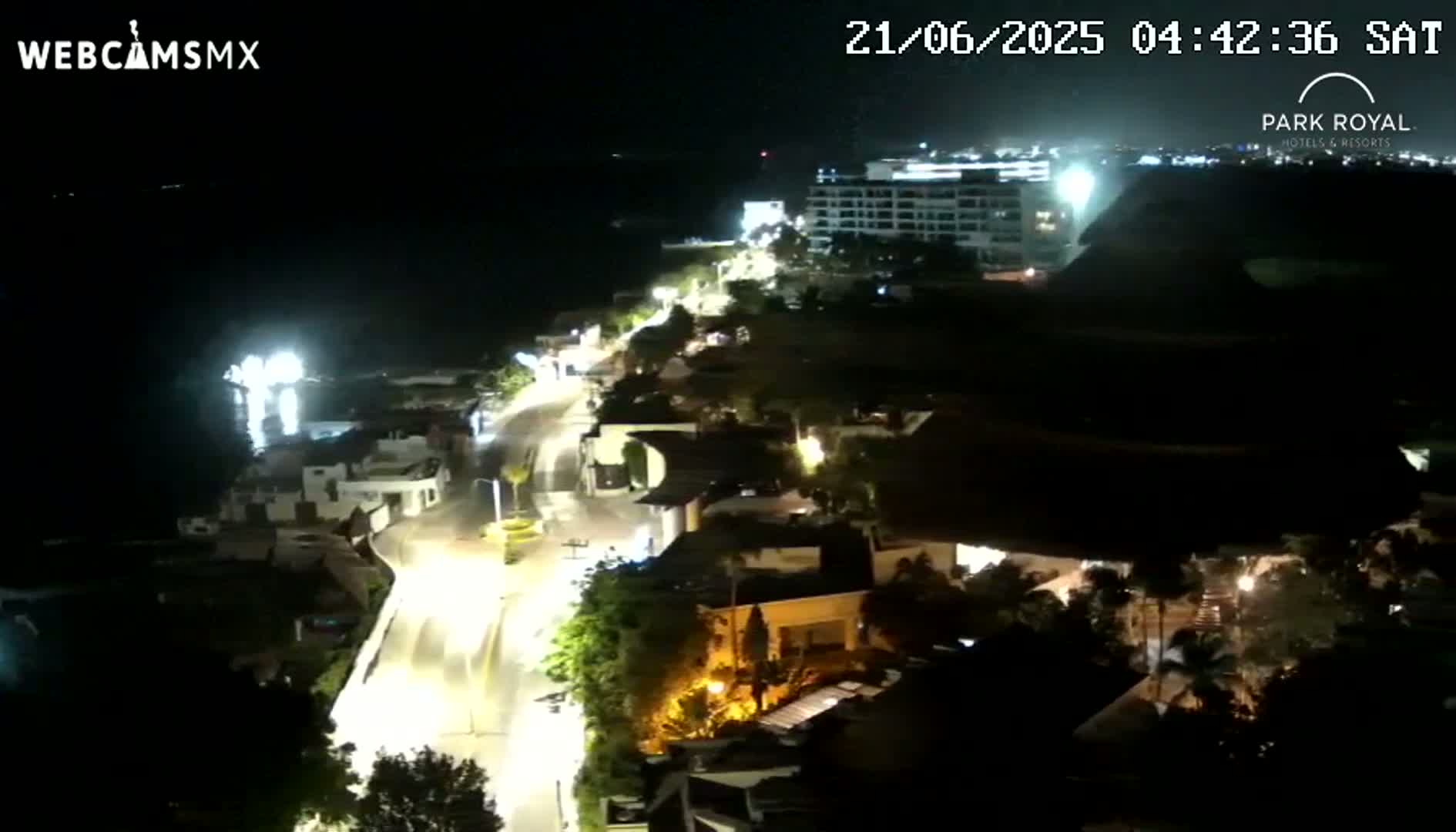 A nighttime, coastal view of a town with a road leading toward a large hotel and the ocean, under clear skies.