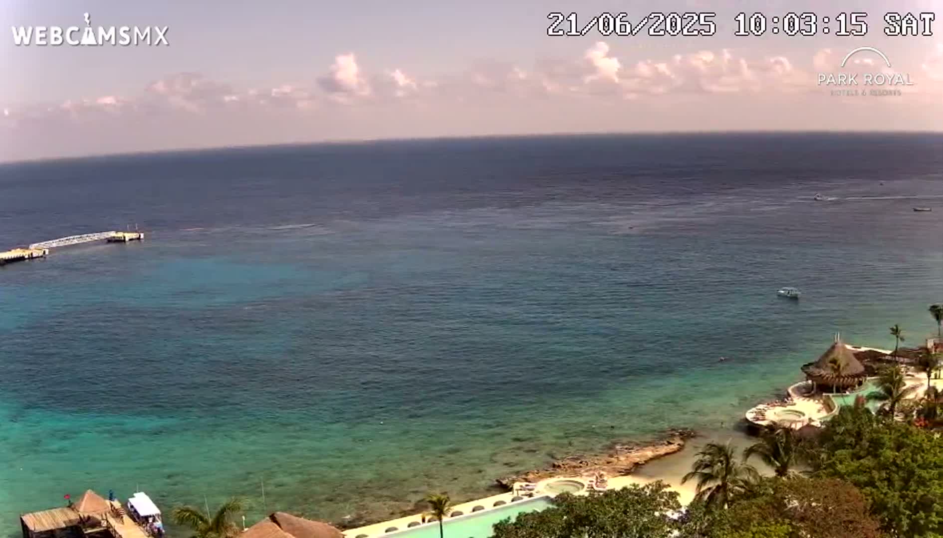 A partly cloudy day shows a tranquil ocean scene with a resort nestled on a rocky shoreline, featuring a swimming pool and thatched-roof structures.