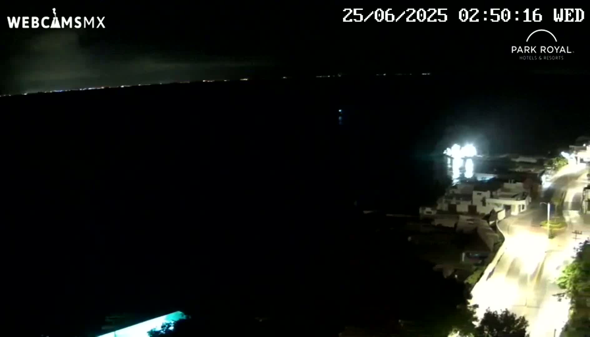 A nighttime coastal scene shows a dark body of water with a faintly lit horizon, a brightly lit road curving along the shoreline, and a few buildings illuminated.