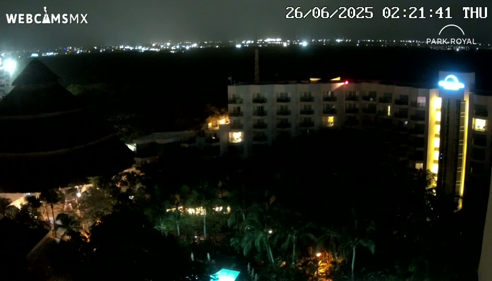 A nighttime view of a resort complex with a lit-up hotel building surrounded by palm trees and a swimming pool, overlooking a city skyline in the distance under a dark sky.
