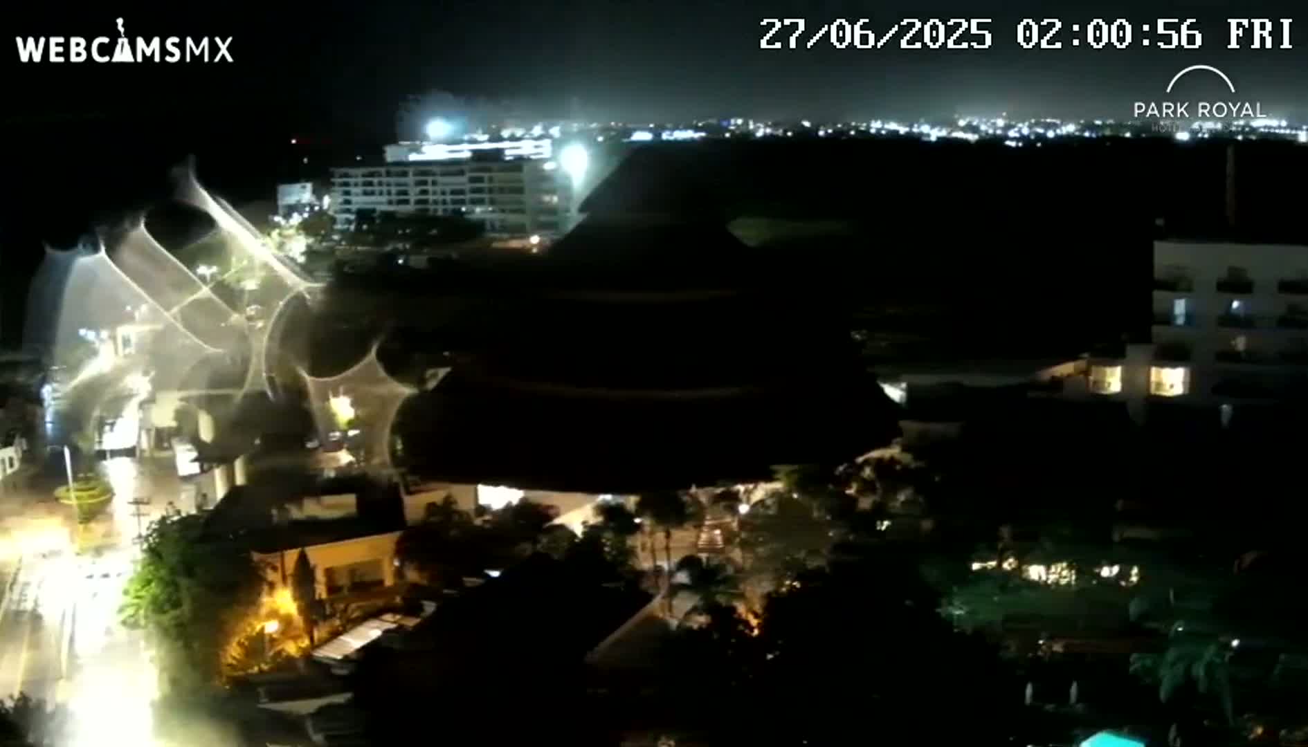 A nighttime aerial view shows a large, illuminated spiderweb-like structure over a city, with buildings and vegetation illuminated by lights.