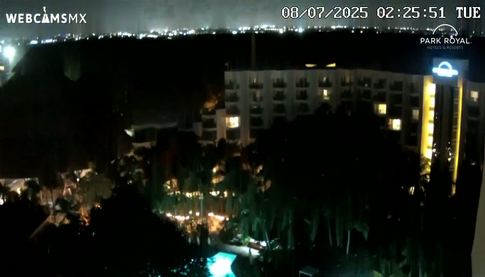 A nighttime, high-angle view shows a large resort complex with illuminated buildings and palm trees surrounding a pool.