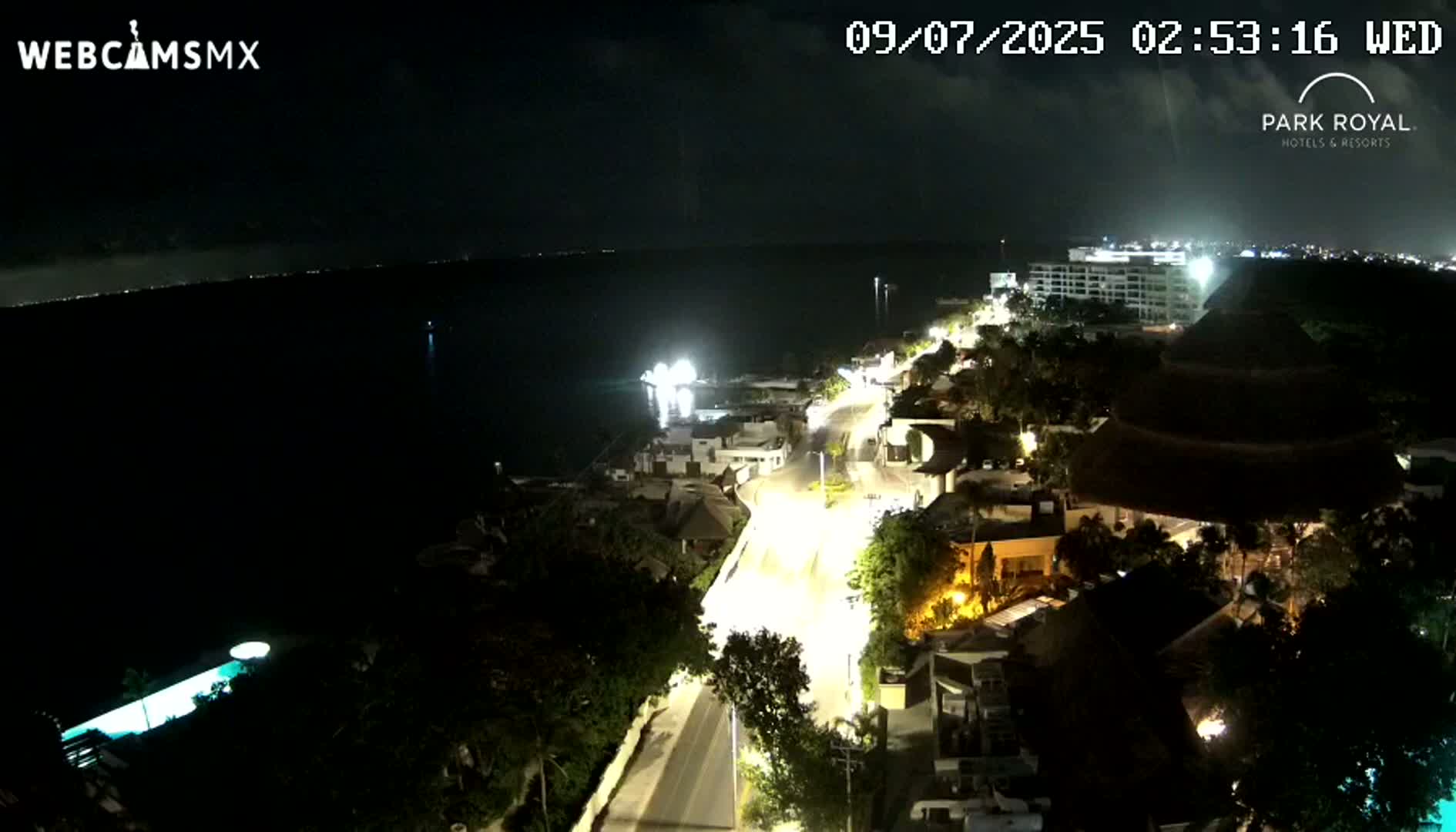 A nighttime aerial view shows a coastal town illuminated with streetlights, next to a dark body of water under a mostly clear sky.
