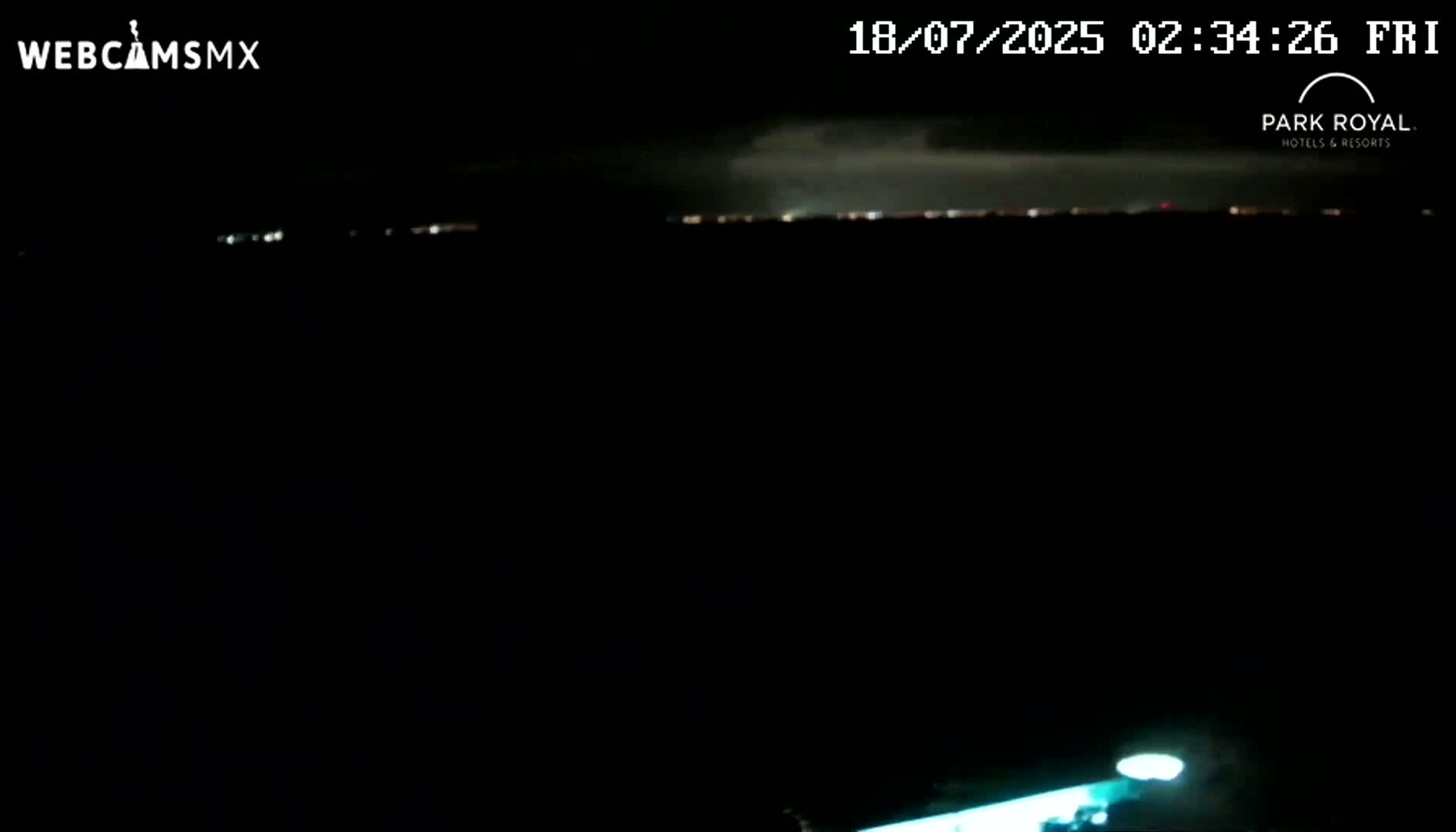 A nighttime, dark scene shows a distant line of lights, possibly a city skyline, under a partly cloudy sky with some lightning visible.