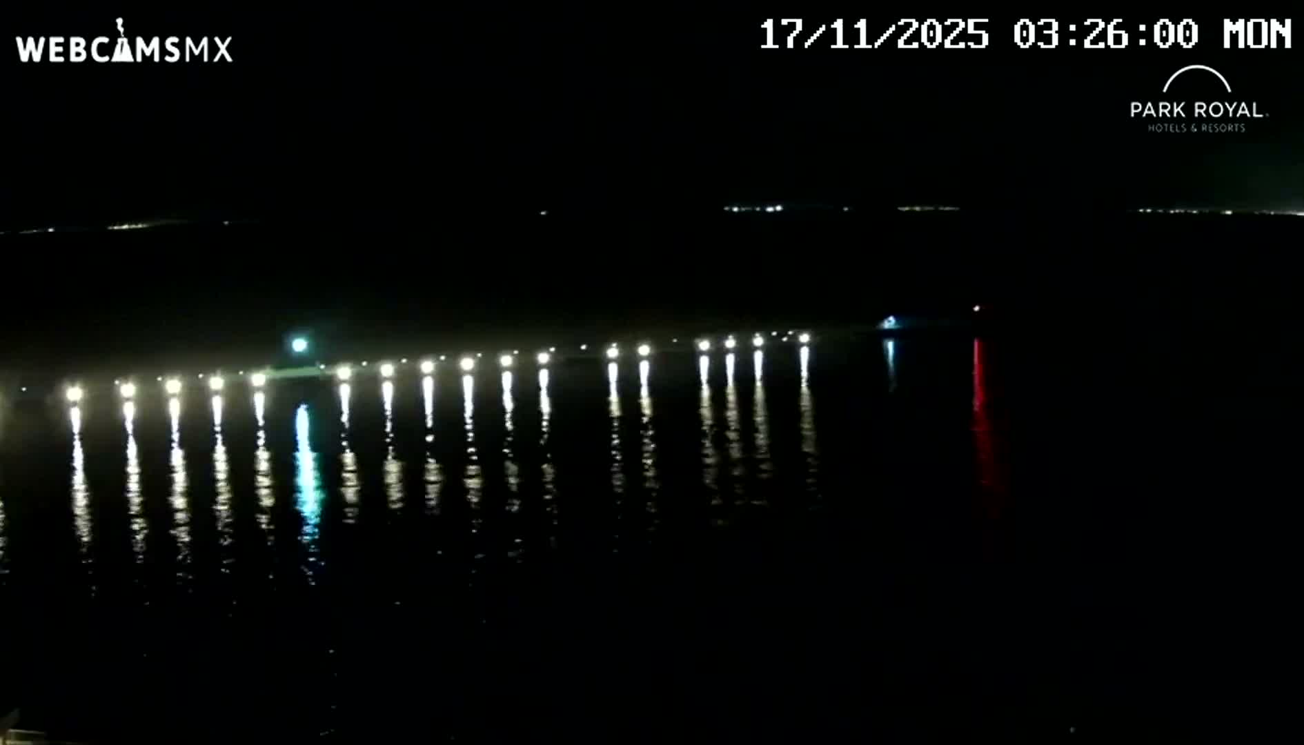 A clear, dark night reveals a long row of illuminated lights stretching across calm water, with their reflections prominent below and a distinct red light visible on the right.